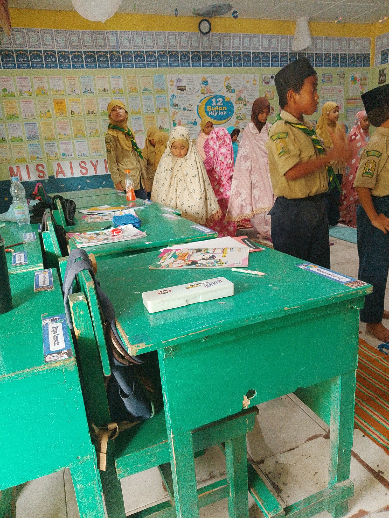 The image depicts a classroom setting, likely an Islamic school, with children in uniform standing and sitting at green wooden desks. The walls are adorned with numerous posters and charts, some with Arabic calligraphy and educational content, suggesting a focus on Islamic studies and general education. In the foreground, a young boy wearing a black cap and a khaki uniform with a green scarf is seen in profile, facing to the right. To his left, a young girl in a pale cream hijab is partially visible. Behind them, other children are also present, some in similar uniforms and headscarves, in various poses suggesting a lesson or activity is underway.
The desks are cluttered with books, papers, a pencil case, and a bottle of water. Labels with names are affixed to some of the desks, indicating assigned seating. The overall environment appears well-lit, with fluorescent lighting and what seems to be a white ceiling fixture. The floor is tiled, and a striped mat is visible in the background, possibly for prayer or seating. The presence of the "MIS AISYI" sign on the wall helps to identify the location.
Based on the available information, this scene is located at MIS Aisyiyah in Tembung, Indonesia. MIS Aisyiyah is an abbreviation for Madrasah Ibtidaiyah Swasta Aisyiyah, which translates to "Private Aisyiyah Elementary School." Tembung is a sub-district within the Deli Serdang Regency in North Sumatra province, Indonesia. The school likely caters to primary-aged children and follows an Islamic curriculum, as evidenced by the posters and the children's attire. The image captures a moment of daily school life, showcasing the educational environment and the students.