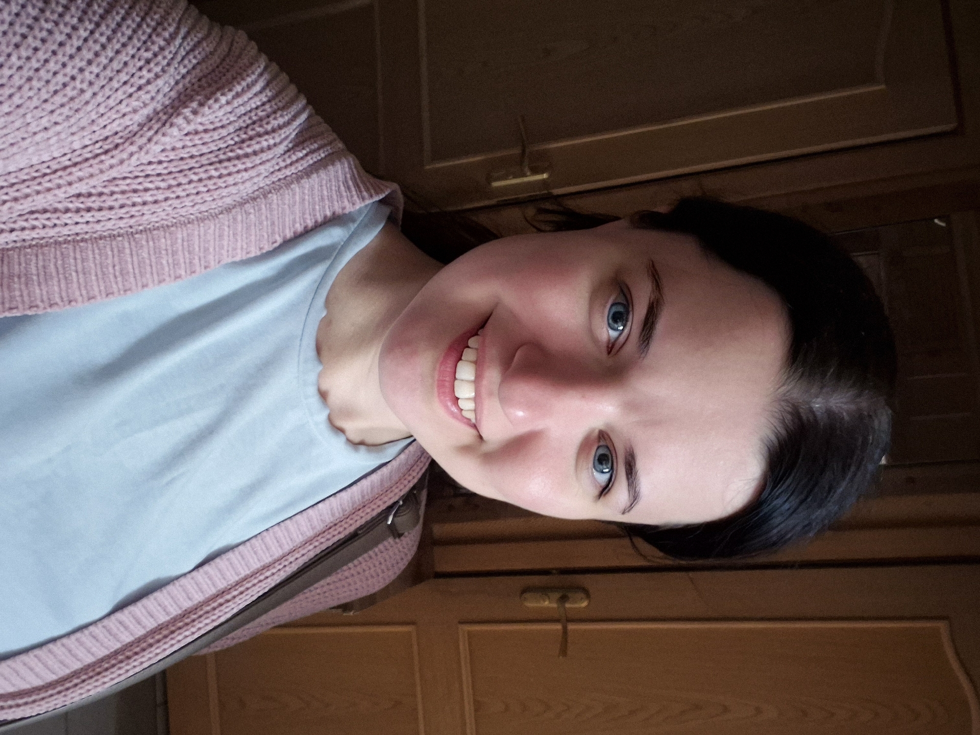 A young woman with dark hair and blue eyes smiles at the camera, taking a selfie. She is wearing a light blue t-shirt and a pink knitted cardigan. Behind her, two wooden doors are visible, one with a brass handle. The lighting suggests an indoor setting, possibly a hallway or room. The image does not contain enough information to determine a specific location within Saint Petersburg, Russia.