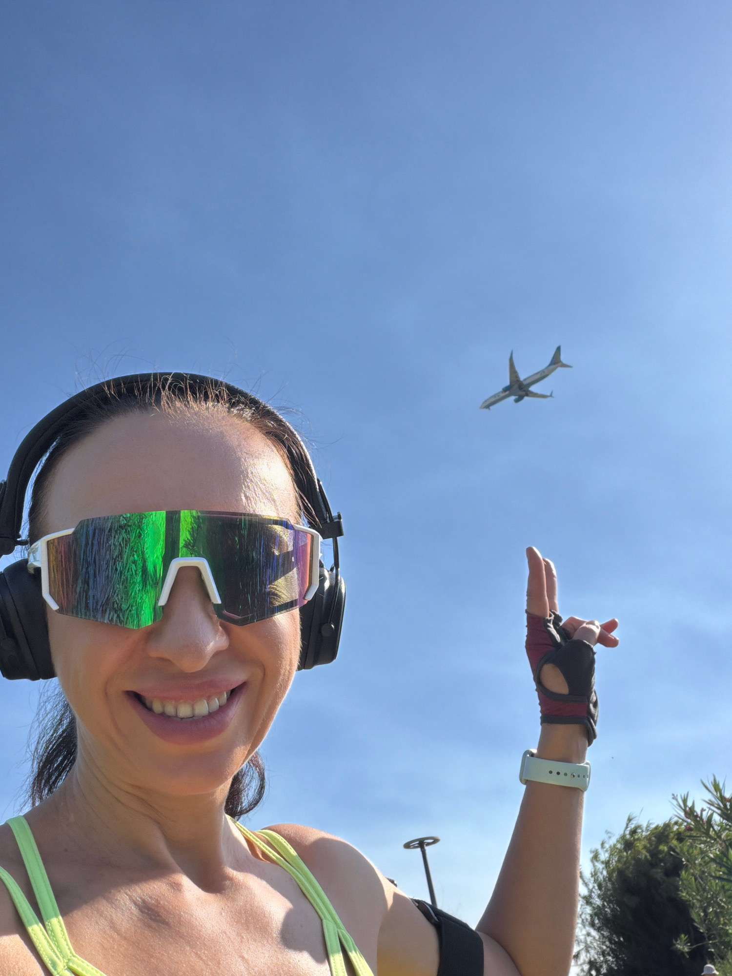 A woman wearing athletic gear, including reflective sunglasses, headphones, and fingerless gloves, smiles at the camera as she extends her right arm upwards, pointing her index finger towards a distant airplane flying in the clear blue sky. The background suggests an outdoor setting, possibly a park or a scenic overlook, with faint hints of greenery and structures, consistent with locations found in Antalya, Turkey.