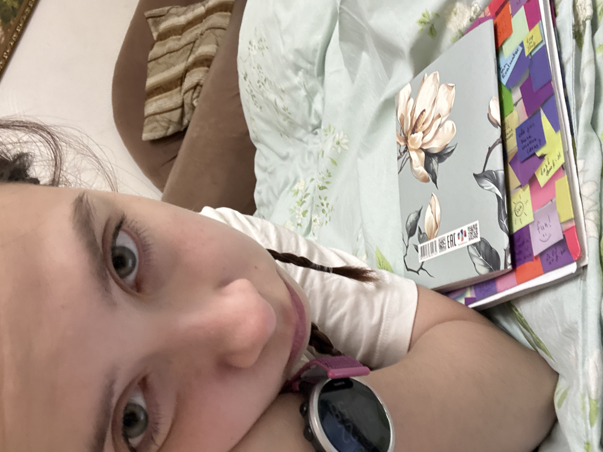 A young person lies on a bed covered with a light green floral patterned blanket. The person has fair skin, grey eyes, and brown hair styled in a braid. They are wearing a white t-shirt and a pink fitness tracker watch on their left wrist. Resting on the blanket near the person's shoulder is a stack of notebooks. The top notebook has a floral design of magnolia blossoms on its cover, and several colorful sticky notes are attached to the sides of the notebooks. A striped pillow or cushion is also visible in the background. There is no information available in the image to confirm the location as the city of Belaya Pashnya, Russia.