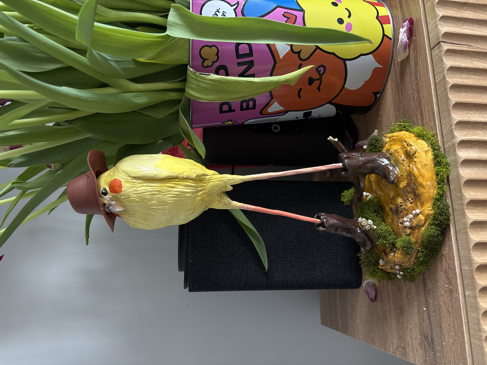 The image presents an indoor setting in Krasnoyarsk, Russia, centered on a stylized avian figurine and a potted plant displayed on a wooden surface. The figurine, approximately 25 cm tall, is a yellow, textured form resembling a chick, featuring orange cheek markings, a small beak, and black eyes. It wears a small brown cowboy hat and stands on long, slender pink legs that terminate in dark brown cowboy boots. These boots are anchored into a small, irregular base composed of a central golden-yellow, textured mound surrounded by green moss-like material dotted with small white floral clusters.
To the left of the figurine, a dark grey rectangular speaker with a woven grille is positioned. To the right, a vibrant pink cylindrical pot holds a plant with numerous upright, elongated green leaves, suggestive of tulips. The pot is decorated with cartoon depictions of an orange fox and a yellow character, alongside partial text in English ("P. BND") and Cyrillic ("Уу..у!"). Several reddish-purple flower petals or buds are visible among the green leaves, and two similar detached petals rest on the wooden surface near the figurine's base and closer to the pot.
The wooden surface exhibits a light brown grain and features a decorative, fluted edge at the front. The background is a plain light grey wall. The scene is evenly lit by ambient light, casting soft shadows.