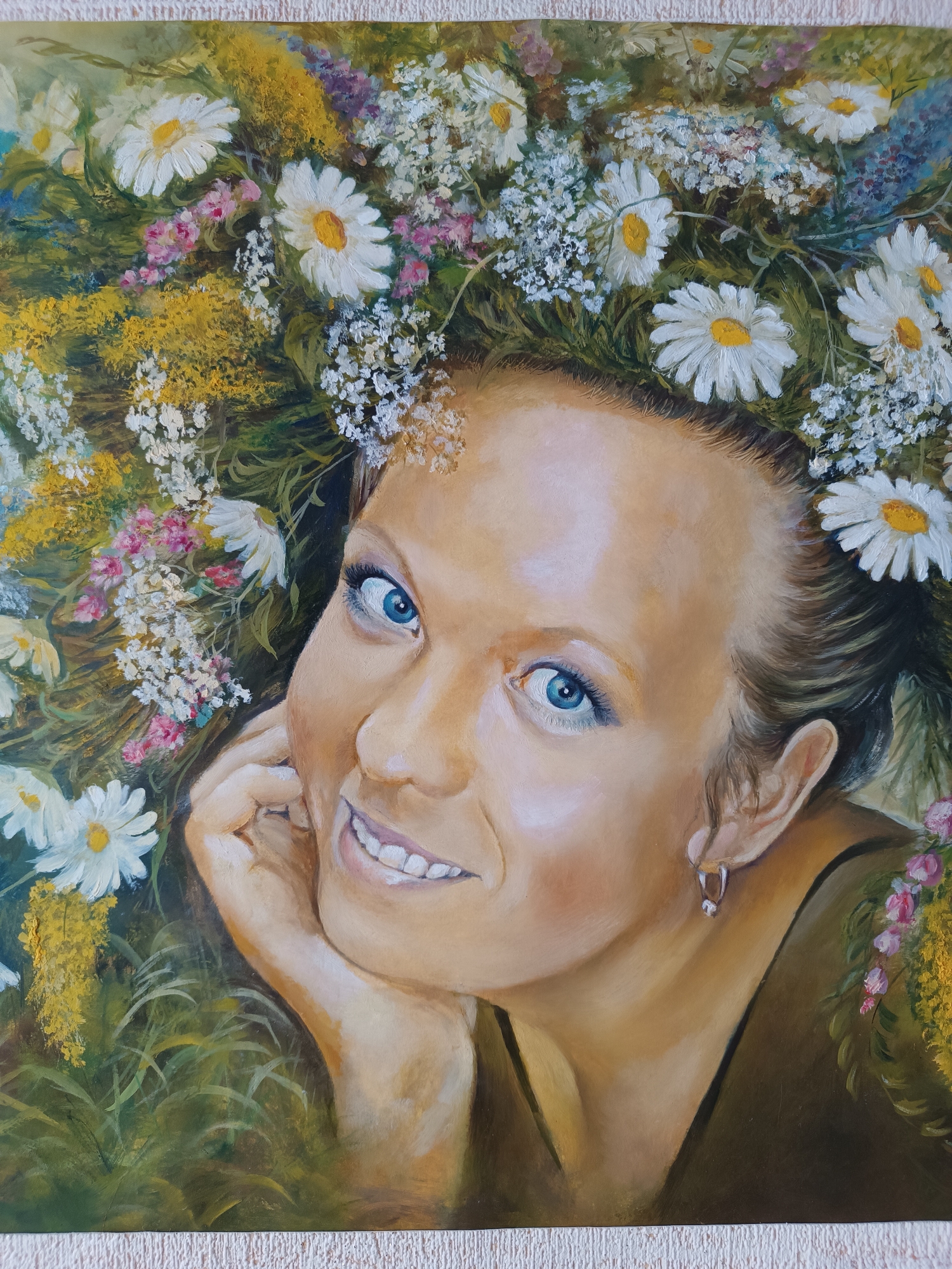 A portrait of a woman wearing a flower crown. The woman has fair skin, blue eyes, and a warm smile. Her hand is resting on her cheek. The crown is composed of white daisies and other small colorful flowers. The background is filled with green and yellow foliage. The location of Пешнигорт, Russia is not discernible from the image.