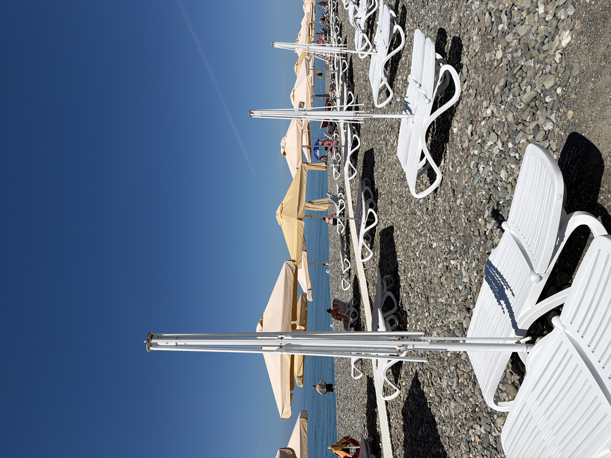 A view of a pebble beach in Sochi, Russia, shows multiple rows of empty white plastic lounge chairs and beige umbrellas. The sea, calm and blue, stretches to the horizon under a clear blue sky. A few people are visible in the distance, some sitting on lounge chairs under umbrellas, while others stand near the water. A red and orange life preserver hangs on a pole to the left of the frame. The foreground is dominated by the white lounge chairs and the pebbly ground.