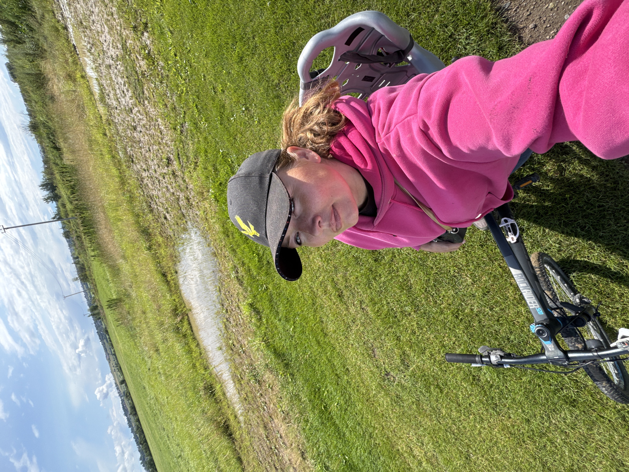 The image is a selfie taken by a woman sitting on a bicycle in a grassy field. The woman is wearing a pink hooded sweatshirt and a black baseball cap with a yellow emblem. She has red marks on her cheeks, likely from sunburn. The bicycle is a mountain bike with the brand name "ROCKRIDER" visible on the frame. In the background, there is a large puddle of water in the field, with more vegetation and a few trees in the distance. The sky is blue with some scattered clouds. The location is identified as the city of Innolovo, Russia.