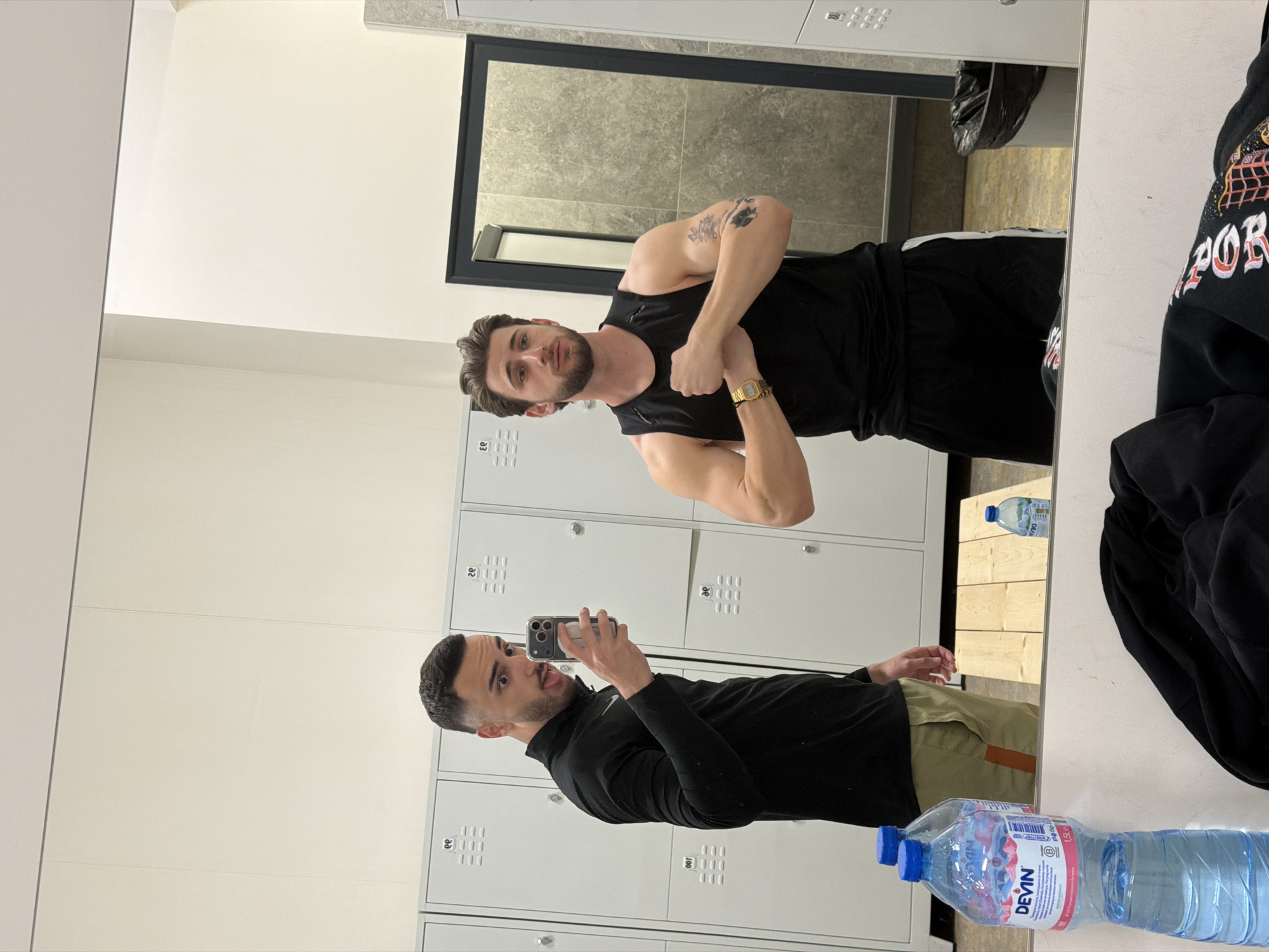 A man is taking a selfie in a gym locker room. He is standing in front of a wall of lockers and holding his phone up to take the photo. His reflection is visible in the mirror, showing him flexing his biceps. The locker room is clean and well-lit. There is a bottle of Devin water visible, and given it is written in Latin script, it is likely a recent edition. The gym may be in Plovdiv, Bulgaria, as Devin is a popular brand of water there.