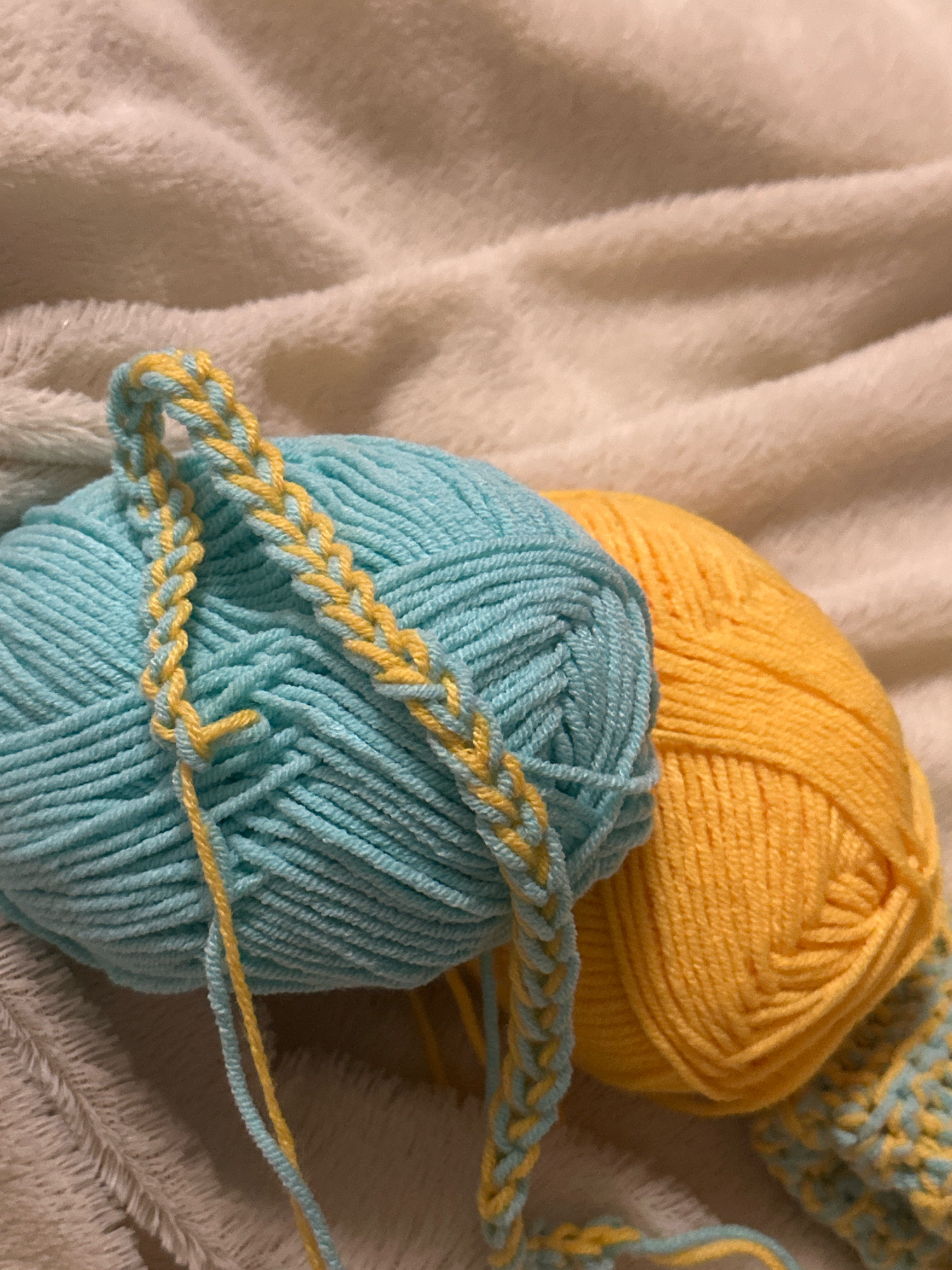 Two balls of yarn, one blue and one yellow, are resting on a plush, cream-colored blanket. A crocheted chain made of blue and yellow yarn is draped over the blue ball of yarn. The scene is indoors. There is no information in the image to suggest a specific location within the city of Gyumri, Armenia.