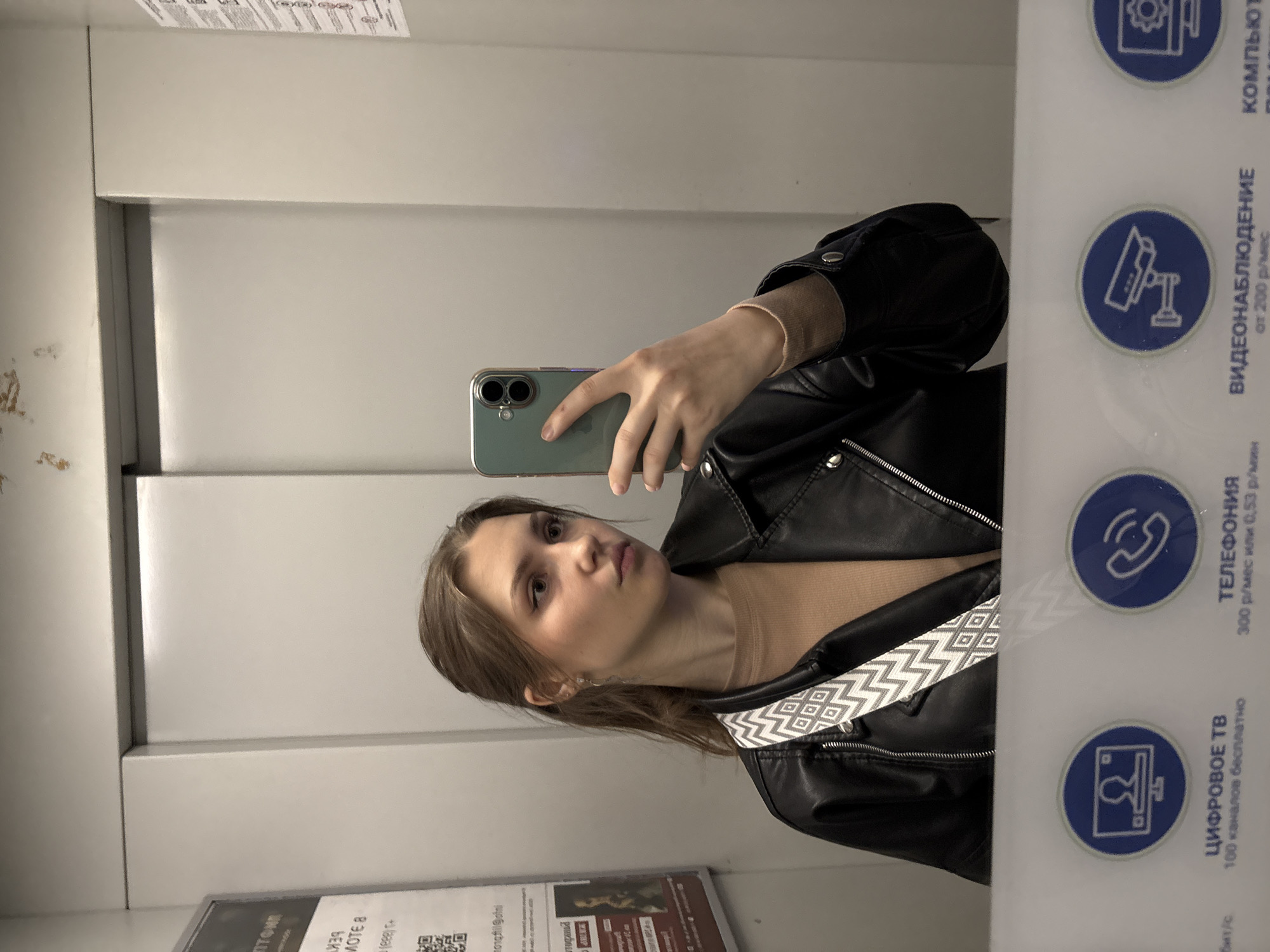A young woman is taking a mirror selfie in what appears to be an elevator or a small room with reflective walls. She holds a green/teal smartphone with a clear case in her right hand, aiming it towards herself. The woman has light brown hair, pulled back into a low ponytail, and a neutral, slightly pouty expression as she looks at the camera. She is wearing a black leather jacket over a light brown, possibly ribbed, top. A patterned strap, with a white and gray geometric design, is visible across her chest. A small earring can be seen in her left ear.
The background consists of light gray or off-white walls, some showing scuff marks on the left side. On the right side of the mirror, a white panel with blue circular icons and Russian text is visible. The icons represent a computer, a CCTV camera, a telephone, and a television.
Visible text includes information about services: "ЦИФРОВОЕ ТВ" (DIGITAL TV) offering "100 каналов бесплатно" (100 channels free); "ТЕЛЕФОНИЯ" (TELEPHONY) at "300 р/мес или 0,53 р/мин" (300 rubles/month or 0.53 rubles/min); and "ВИДЕОНАБЛЮДЕНИЕ" (VIDEO SURVEILLANCE) "от 200 р/мес" (from 200 rubles/month). A partial word "компьют" (likely for computer) is also seen. Other partial text like "Птол", "MOTE 8", and "бит/с" appears on posters or labels. The presence of Russian text aligns with the given location context of Saint Petersburg, Russia. The image is taken indoors, with no indication of time of day or weather.