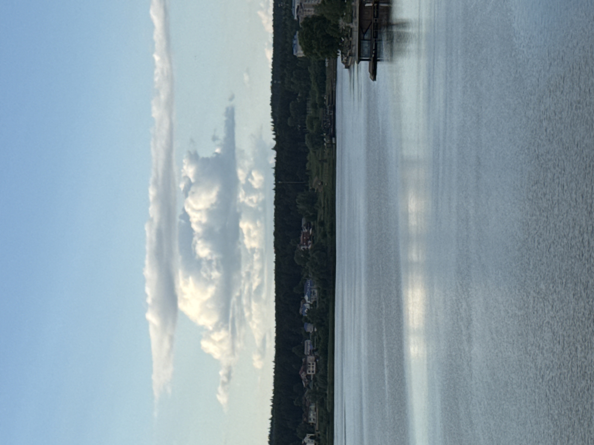 This image, when viewed horizontally, captures a serene landscape featuring a body of water, a tree-lined shore, and a blue sky with clouds.
A wide expanse of calm water, likely a lake or river, dominates the lower and right portions of the scene. Its surface shows gentle reflections of the sky and light, appearing speckled and slightly textured. Along the far bank, a dense, dark green forest forms a horizontal band, stretching across the middle of the image. Within this treeline, several houses or buildings of varying sizes and colors (including white, red, and darker tones) are visible. On the right side of the shoreline, a structure, possibly a boathouse or pier, extends slightly into the water.
The upper left portion of the image is filled with a bright blue sky, adorned with scattered white cumulus clouds. One long, elongated cloud formation is particularly prominent. The lighting suggests late afternoon or early evening, creating a soft and tranquil atmosphere. No specific activity or individuals are visible. The location context for this peaceful scene is Votkinsk, Russia. No readable text is discernible.