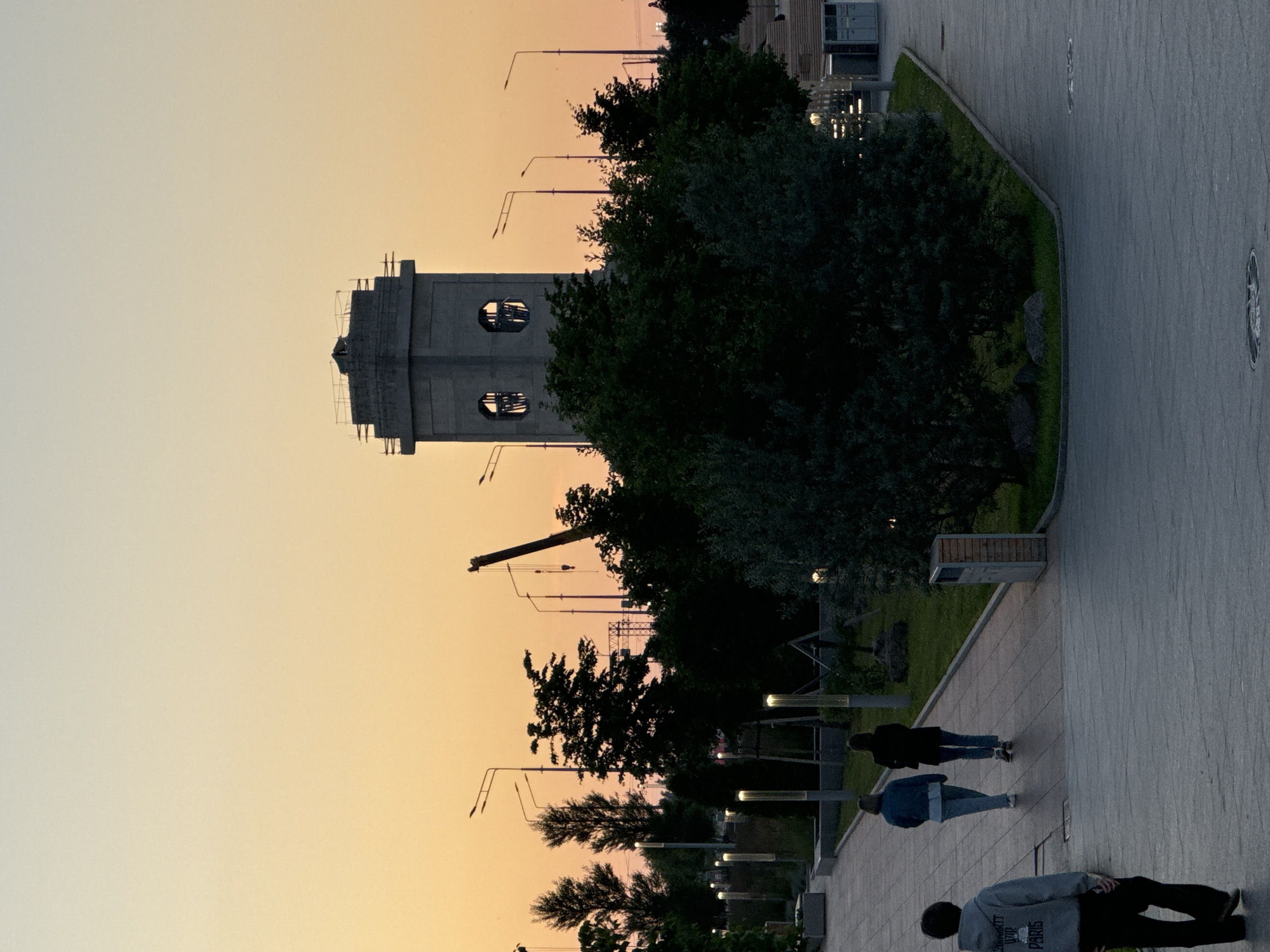In Sinyavino, Russia, a dusk scene unfolds in a park-like setting. A tall, unfinished concrete tower with two round openings dominates the background, appearing under construction with scaffolding. In the middle ground, a group of trees form a dense silhouette against the warm orange and pale yellow gradient of the twilight sky. A construction crane is visible behind the trees. Several lampposts line a pathway in the mid-ground, some illuminated. Three individuals are present: one person in a grey hoodie and dark pants walks away from the viewer on a paved walkway in the foreground. Further down the walkway, two individuals, one in a blue jacket and light pants and the other in a dark jacket and dark pants, walk in the same direction. The ground is paved, and there are grassy areas and some large rocks scattered within the park. A trash receptacle is situated near the pathway.