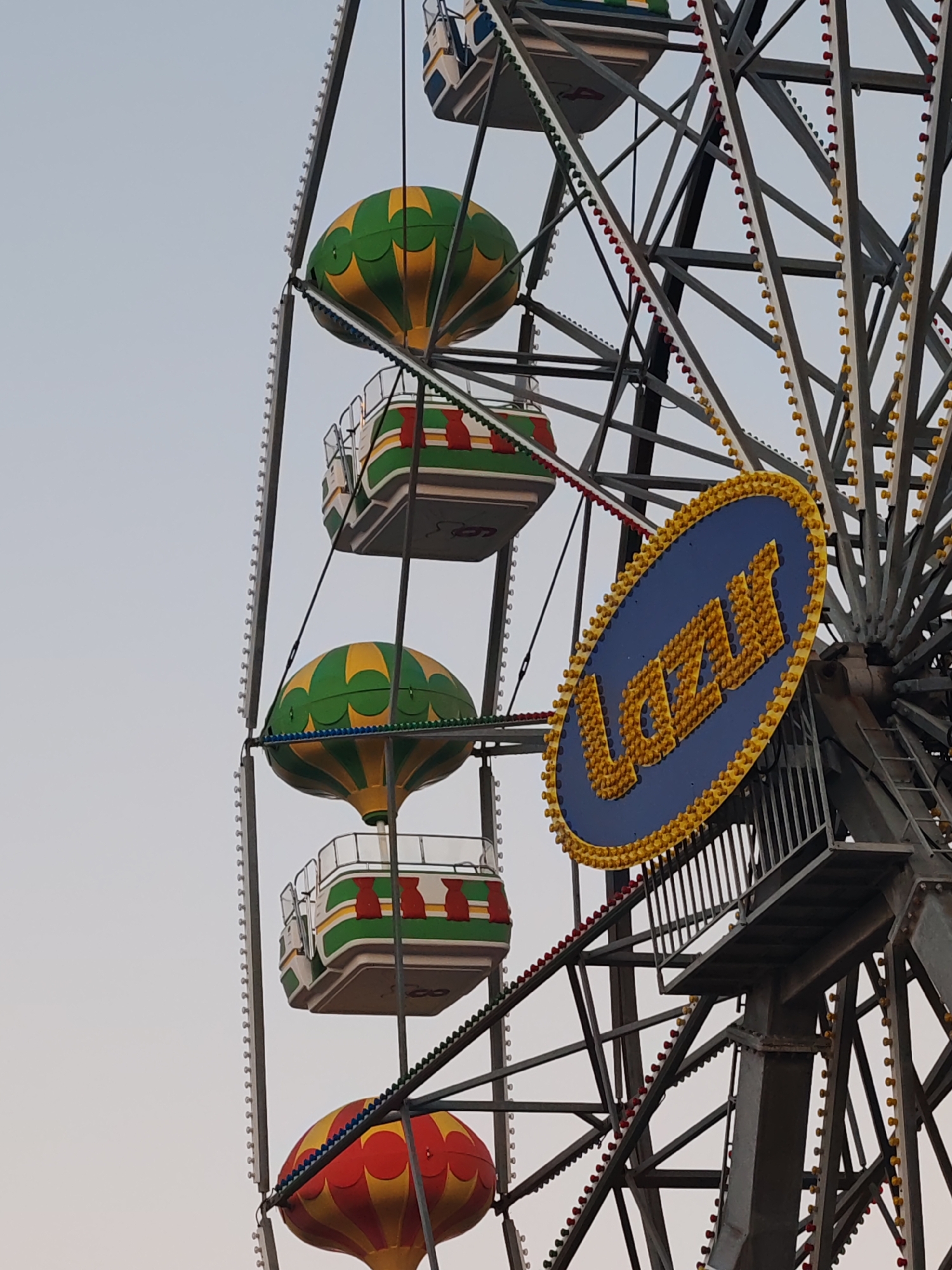The image captures a partial view of a Ferris wheel in Nesebar, Bulgaria. The wheel's design incorporates balloon-shaped gondolas in alternating color patterns, including red and yellow, and green and yellow. The gondolas are attached to the wheel's structure via metal supports. The Ferris wheel has a large, oval-shaped sign at its center, displaying the word "LAZY" in large, yellow letters against a blue background. The sign is bordered with lights. The wheel's metal framework is visible, with lines of lights running along various structural elements. The sky visible in the background is pale, suggesting either dawn or dusk.