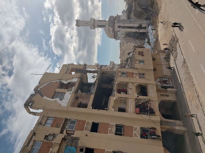 A building ravaged by war sits beside a mosque. The structure's facade is crumbling, with visible signs of fire damage and destruction of inner portions. Garments hang from the window sills, adding a poignant human element. The mosque in the background also shows damage, its dome and minaret bearing the scars of conflict. A road lies adjacent to the building, where few figures are seen walking, juxtaposing normalcy against devastation. The sky is partly cloudy.