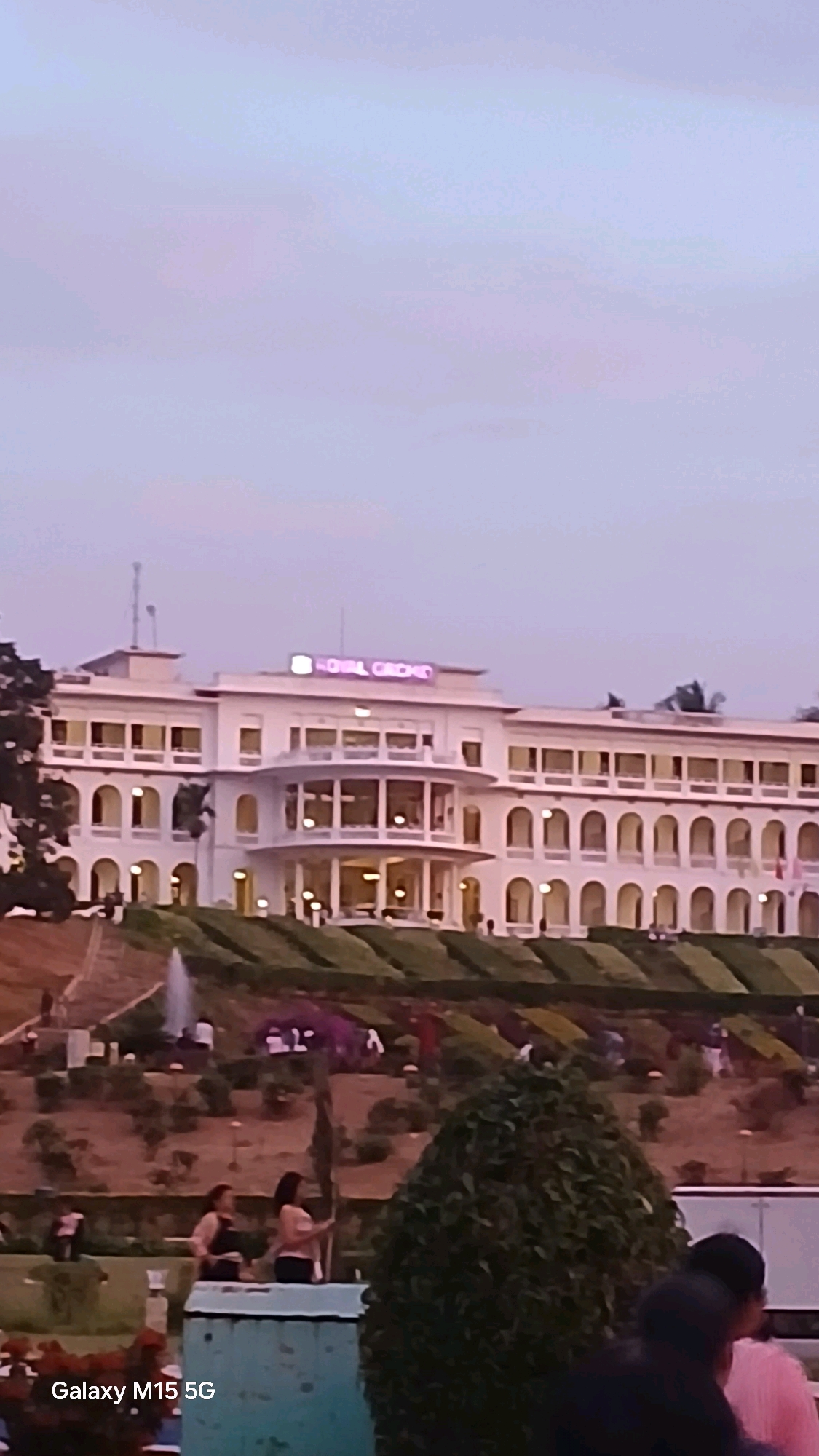 The image depicts the exterior of the Royal Orchid hotel in Krishnarajasagara, India, during twilight. The hotel is a multi-story white building with a prominent curved glass-enclosed section on the lower floors. Illuminated signage on the rooftop reads "ROYAL ORCHID". In front of the hotel, there is a terraced garden with manicured green hedges and several fountains. In the foreground, two women are visible, one holding a flag. The sky is a soft gradient of pale blue and pink, indicating the end of the day. A portion of a tree is also visible in the lower right corner. The bottom left corner of the image contains the text "Galaxy M15 5G".