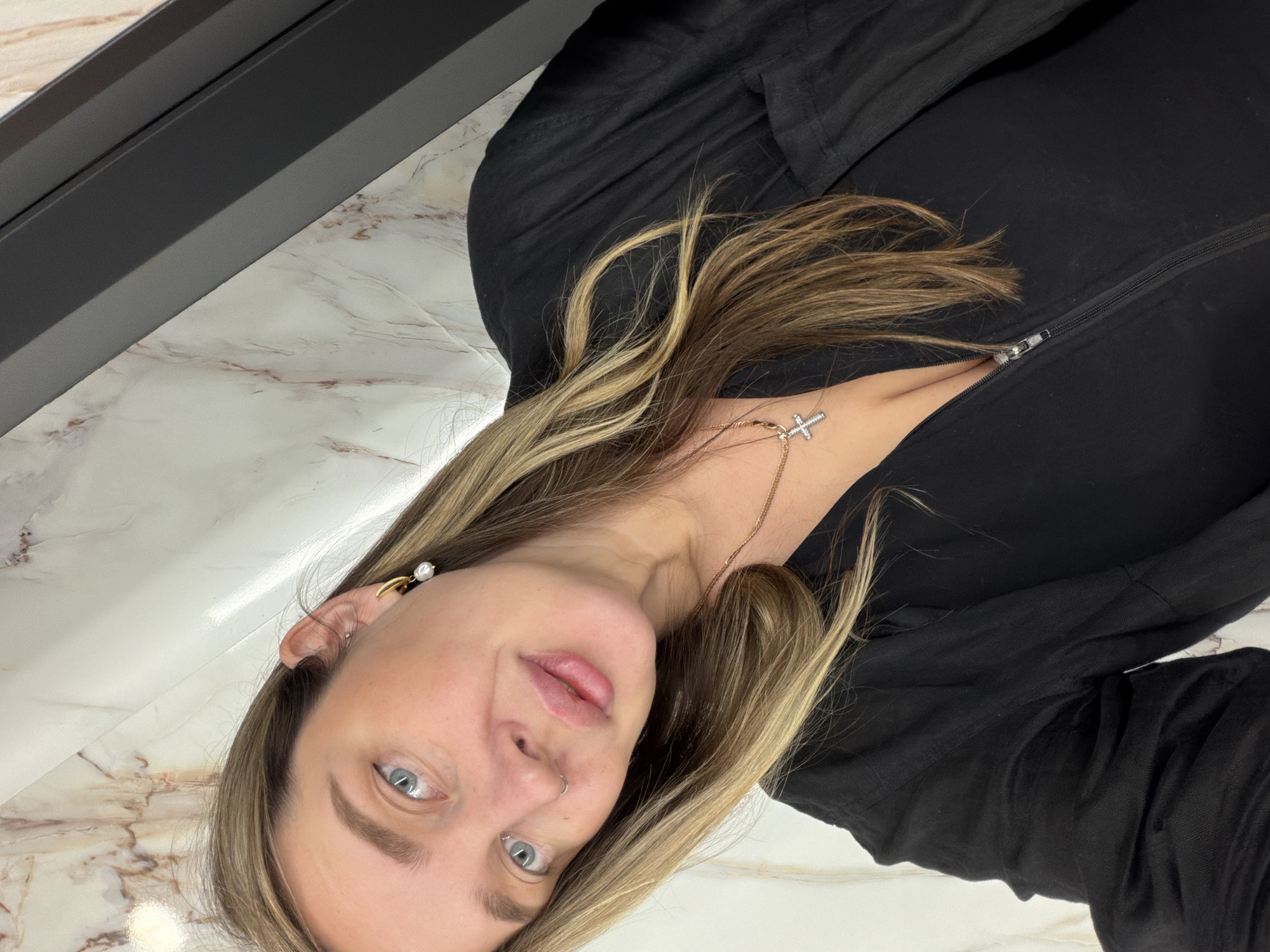 A woman with light brown and blonde hair, blue eyes, and a nose piercing is facing the camera. She is wearing a black zippered jacket and a necklace with a cross pendant. Her ear is visible, showing a gold earring with a pearl accent. The background consists of a white and brown marble surface, and a black object is visible in the upper left corner. There is no indication of the location being Moscow, Russia.