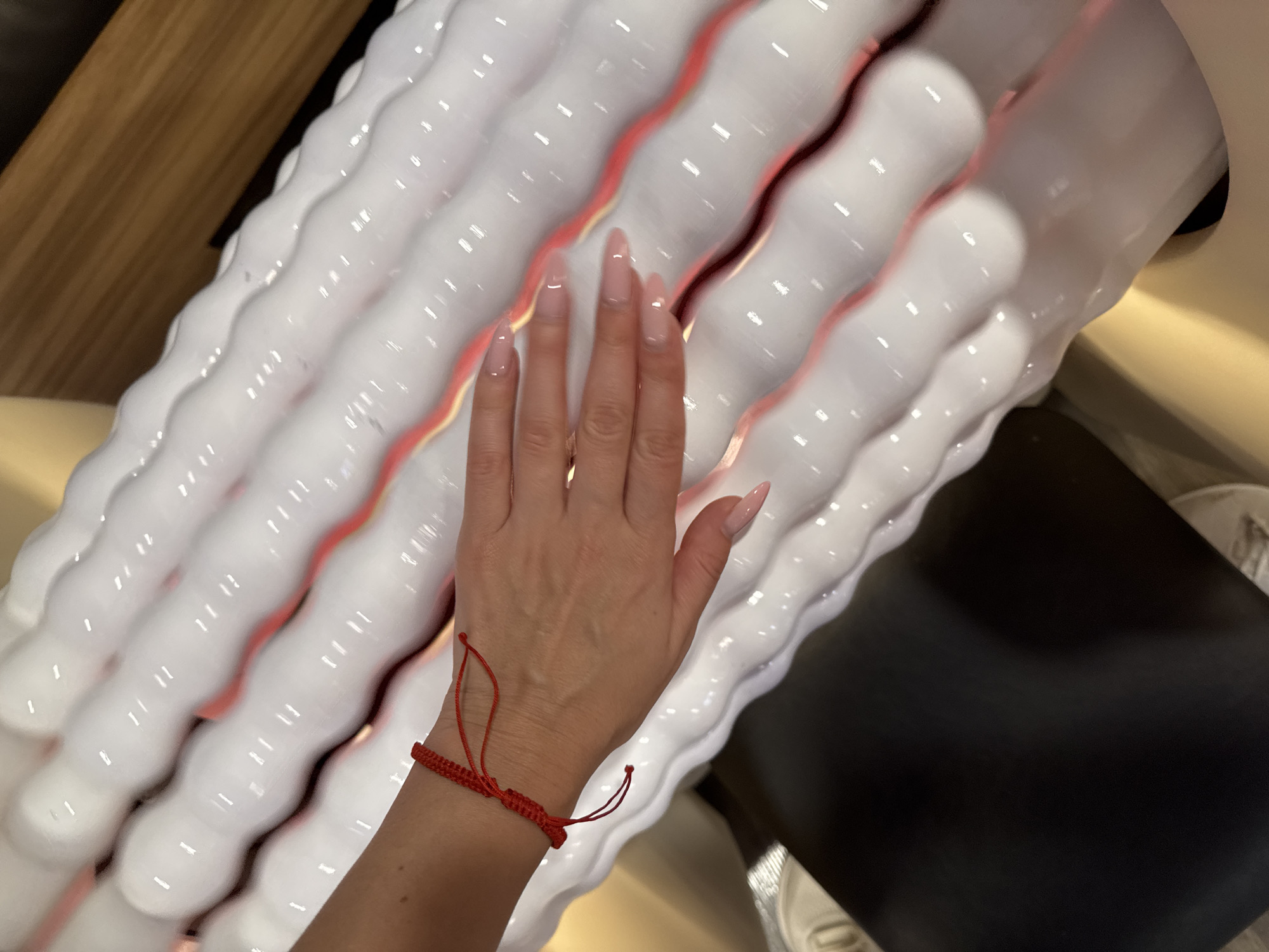 A human left hand, adorned with a red string bracelet on the wrist, rests palm-down on a piece of specialized equipment. The equipment features multiple parallel, wavy, white glossy rollers or tubes, which appear to be internally lit, emitting a soft red glow from the indentations between them. The hand's fingernails are long, almond-shaped, and finished with a light pink or nude polish. In the background, to the upper right, a blurred brown, possibly wooden surface is visible. A dark, matte, black surface occupies the lower-left portion of the frame, and a light beige or tan structure supports the white rollers. The scene is indoors, in the city of Unknown, Unknown.