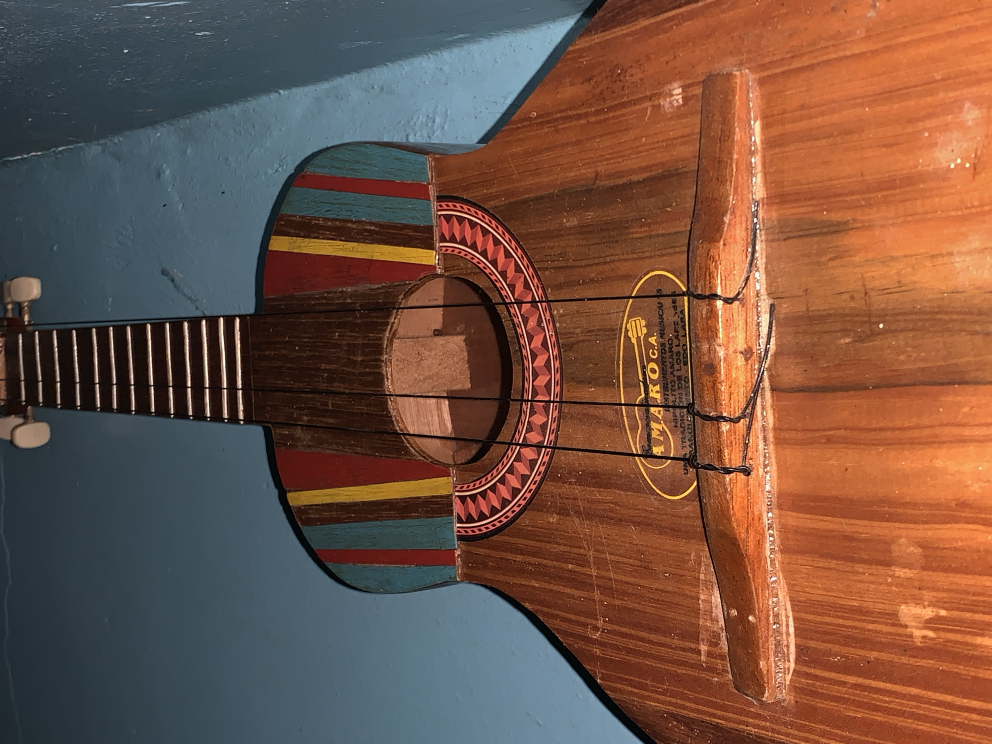 A rustic, four-stringed musical instrument, resembling a Venezuelan cuatro, is depicted leaning vertically against a textured, light blue wall. The instrument features a wooden body with a visible grain, suggesting a handcrafted origin. The upper bout and neck joint are adorned with hand-painted stripes in blue, yellow, and red. A decorative rosette with a geometric red and white pattern encircles the soundhole.
The instrument shows signs of wear and use, including scuffs and scratches on its wooden surface and chipped paint on the colored stripes. Four dark strings are visible, stretching from the partially seen headstock to a simple, wooden bridge that appears to be held in place by the tension of the strings and possibly some adhesive or sealant. A yellow oval-shaped label is affixed to the instrument's body, near the soundhole.
The text on the label, partially captured by OCR, reads: "AMARO C.A.", "INSTRUMENTOS MUSICAIS" (Musical Instruments), and "HIPOLITO AMARO". The last lines, though distorted by OCR as "TRADICION DE LOS LAPZ BES" and "TO-EDO, LARAL", likely refer to "TRADICIÓN DE LOS LLANOS" (tradition of the plains, a region in Venezuela) and "ESTADO LARA" (Lara State), respectively. This, combined with the location context of Santa Ana, Venezuela, strongly suggests the instrument is a traditional Venezuelan cuatro, possibly made by Hipolito Amaro in Lara State, a region known for its musical heritage. No activity is taking place; the instrument is stationary. The lighting suggests an indoor setting with warmth on the instrument's surface.