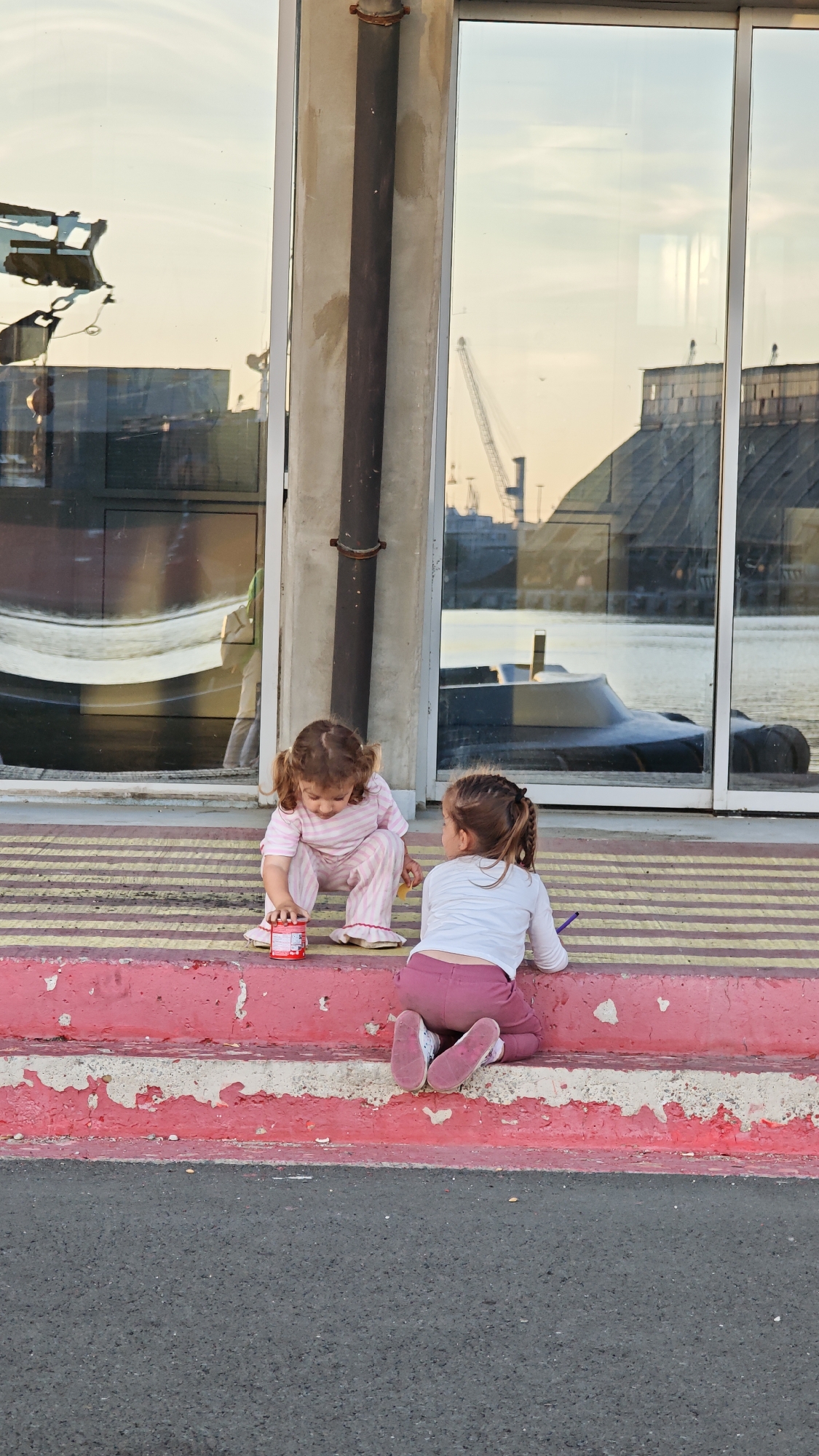 Two young girls are sitting on the edge of a curb in Burgas, Bulgaria. One girl is wearing pink and white striped pajamas and is playing with a red jar. The other girl is wearing a white long-sleeve shirt and pink pants. The curb is painted with alternating horizontal stripes of yellow and pink. Behind them is a building with large windows reflecting the Burgas port. A metal pipe runs vertically along the building’s exterior. The street is asphalt.