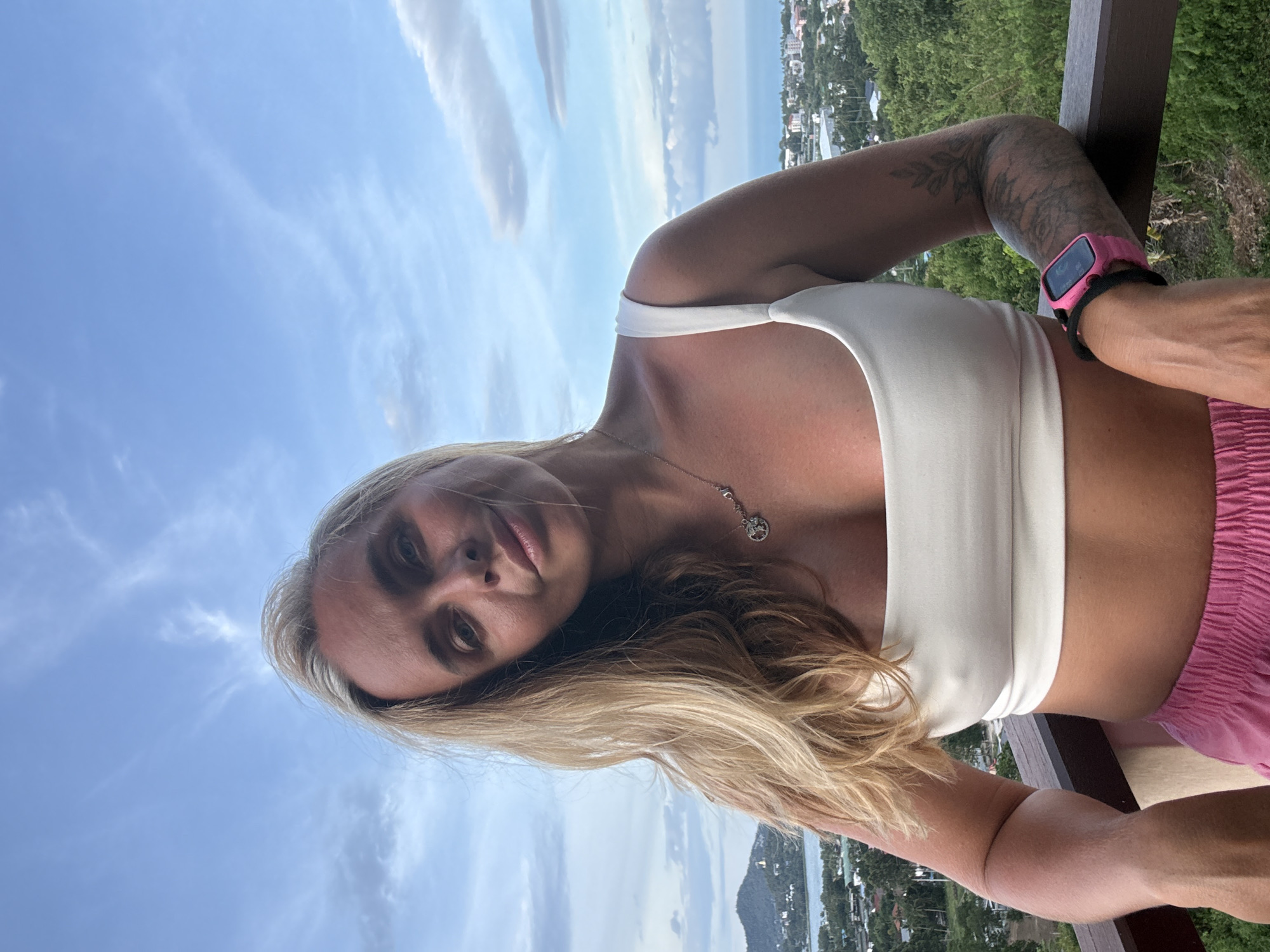 A blonde woman stands on a balcony overlooking a city and ocean view. She is wearing a white crop top and pink shorts. Her hair is long and wavy, and she has a necklace with a circular pendant. She is also wearing a pink smartwatch on her left wrist. The background shows a city with buildings and trees, and the ocean in the distance. The sky is partly cloudy. The location is identified as Koh Samui District, Thailand.