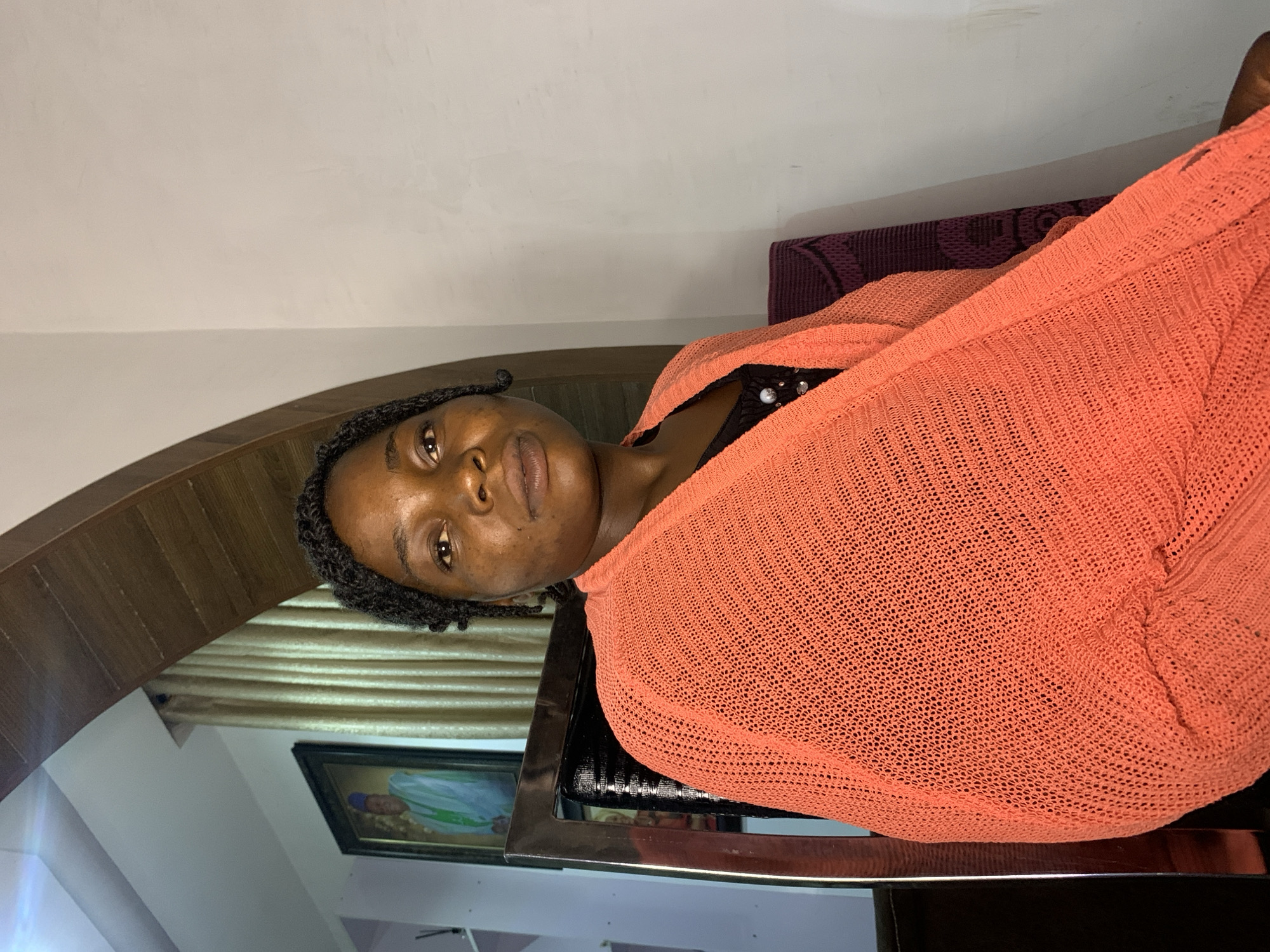 A medium shot captures a Black woman seated indoors, looking towards the camera. She has dark skin, dark eyes, and her dark hair is styled in braids, with some strands framing her face. She wears a black top adorned with embellishments and a vibrant orange knit cardigan draped over her shoulders. Her gaze is direct, and her lips are slightly parted. Behind her, a framed portrait of a man in traditional attire hangs on the wall, partially obscured by a draped curtain. The woman is seated in a chair with a dark, possibly leather, seat and a polished metal frame. The environment appears to be a living room in Jos, Nigeria, indicated by the presence of a portrait and the style of the home decor. The lighting is somewhat dim, with light sources casting shadows.
