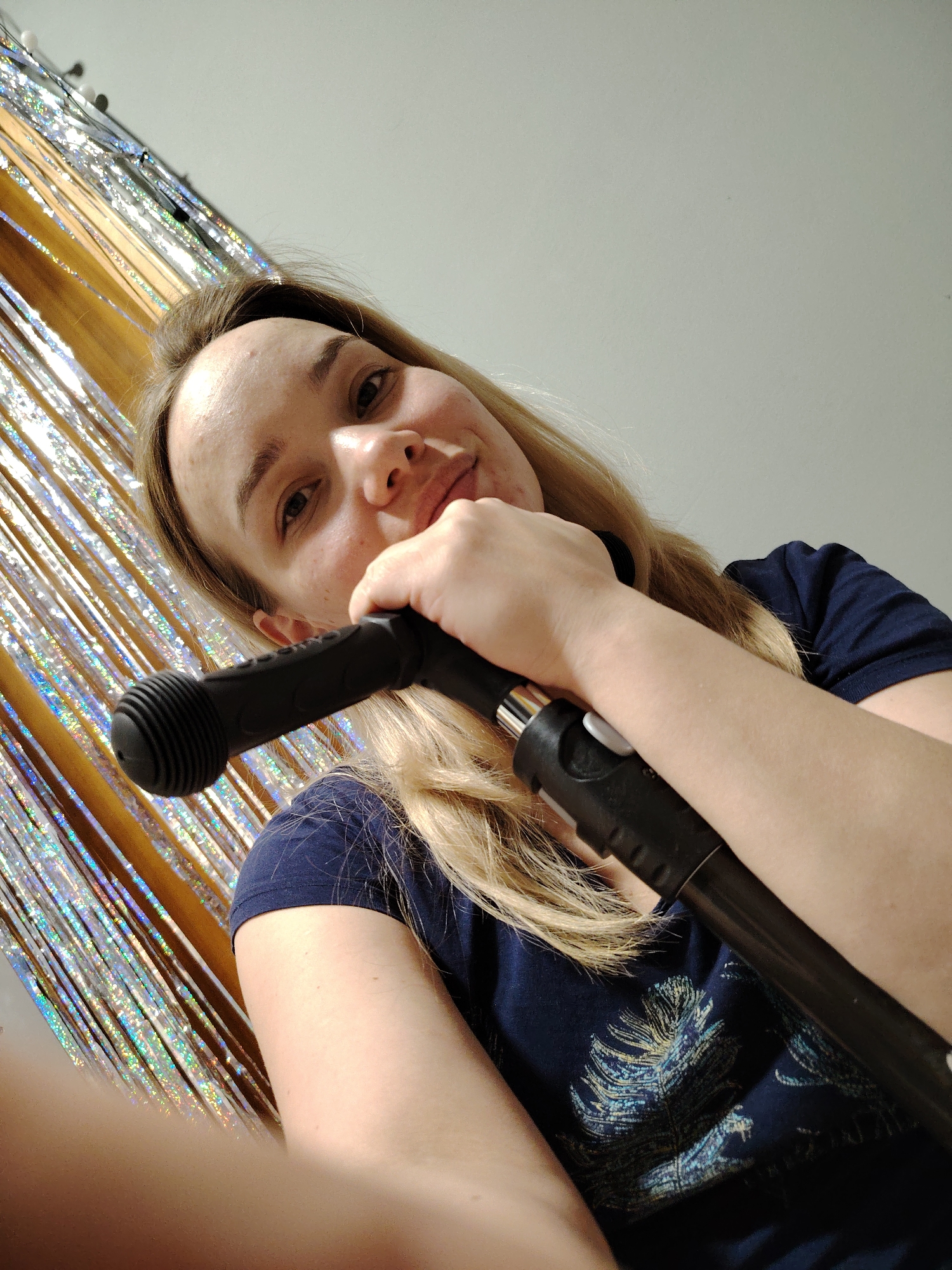 A woman is holding a black microphone stand, with her hand resting on the microphone head. She is smiling and looking at the camera. She has long blonde hair and is wearing a dark blue t-shirt with a feather design. Behind her is a shiny, silver tinsel-like decoration. The background includes a plain white wall. The location is Марковский, Russia.
