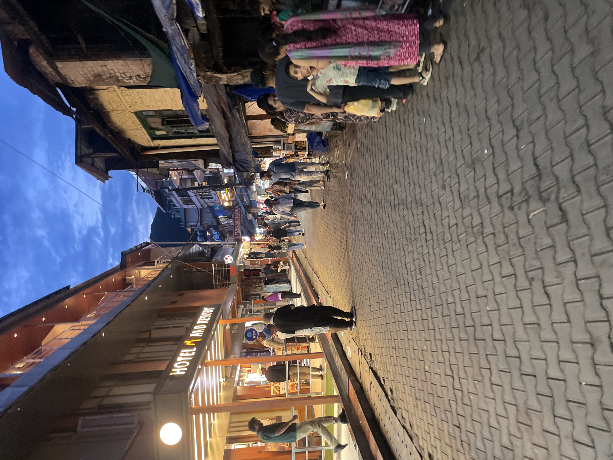 The image depicts a street scene in Dharamshala, India, likely on the evening of a bustling day. The street is paved with interlocking grey bricks and lined with multi-story buildings on both sides. On the left, a hotel named "HOTEL M AND RESORT" is prominently displayed with illuminated signage. Several people are walking on the street or standing near the hotel entrance.
On the right side of the street, there are shops and stalls with awnings and tarps providing shade. A group of people, including a young boy, a woman in a pink and green patterned garment, and others, are standing in a line or near the stalls. Some individuals are casually dressed, while others are in traditional Indian attire. The sky above is a twilight blue with some clouds, suggesting it is either dusk or dawn. The overall atmosphere appears to be lively, with evidence of commercial activity and pedestrian traffic in a densely built urban environment.