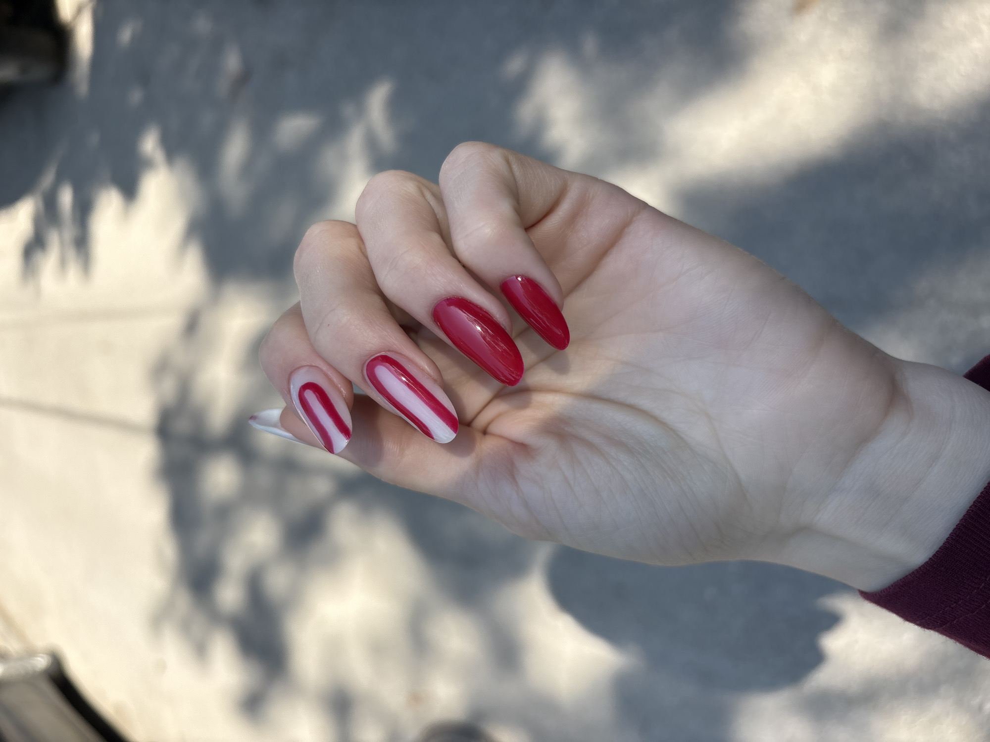 A close-up photograph showcases a hand with long, coffin-shaped nails. Two nails are painted solid red, while the other two feature a white base with a single, thick red stripe running diagonally. The hand is held against an outdoor backdrop with dappled sunlight and shadows cast on a concrete surface. The edge of a burgundy-colored sleeve is visible in the lower right corner. The location, Kragujevac, Serbia, is not visually discernible from the image alone.