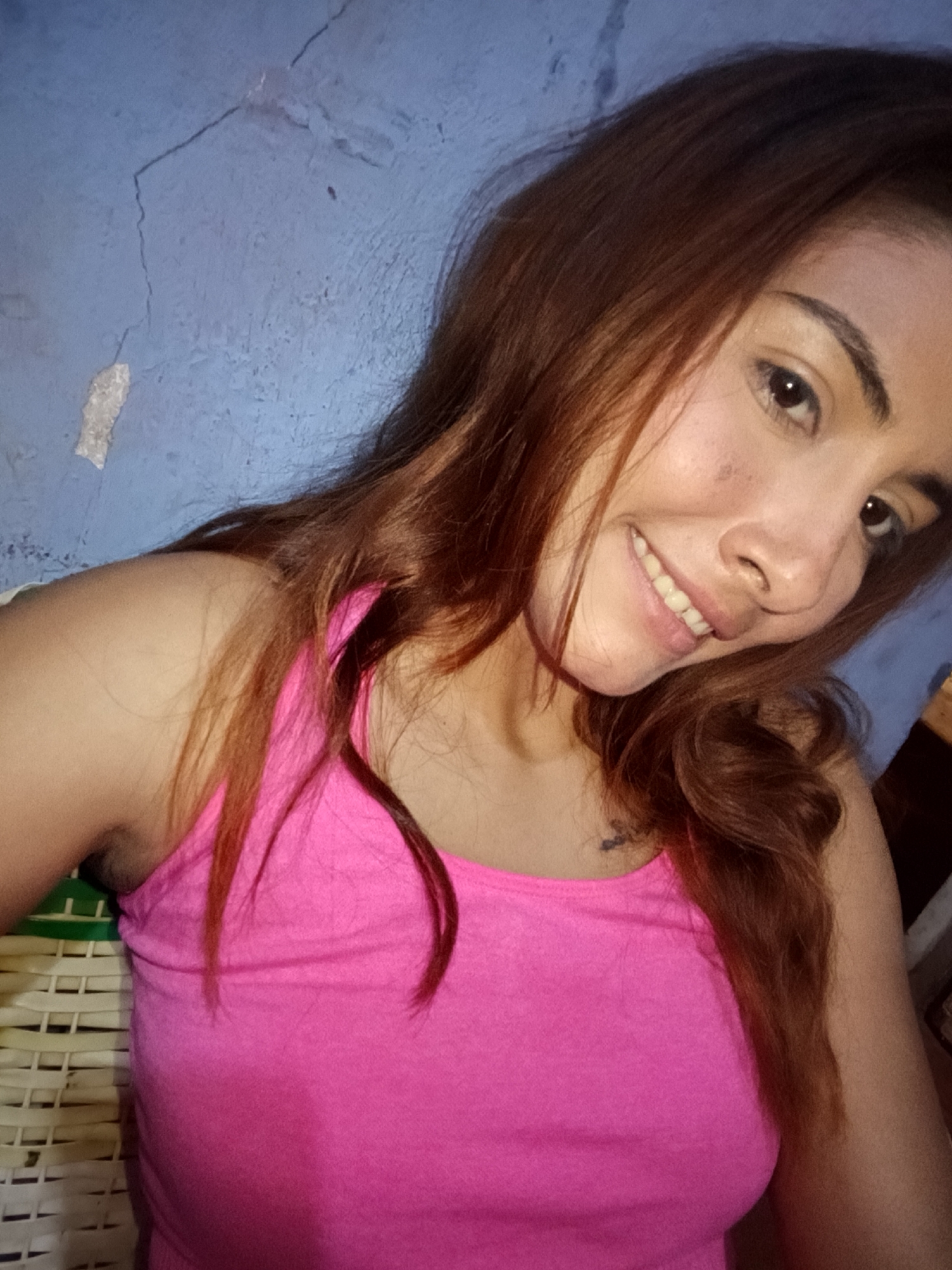 The image shows a close-up, waist-up selfie of a young woman with long, reddish-brown hair, smiling. She is wearing a bright pink tank top. Her skin is light-toned, and she has dark eyes and eyebrows. The background is a textured blue wall with some visible cracks and peeling paint. To the left, a portion of a woven white and green chair or furniture item is visible. The overall lighting suggests an indoor setting. There are no clear indicators of the location being specifically in Juan Griego, Venezuela, beyond the provided context.
