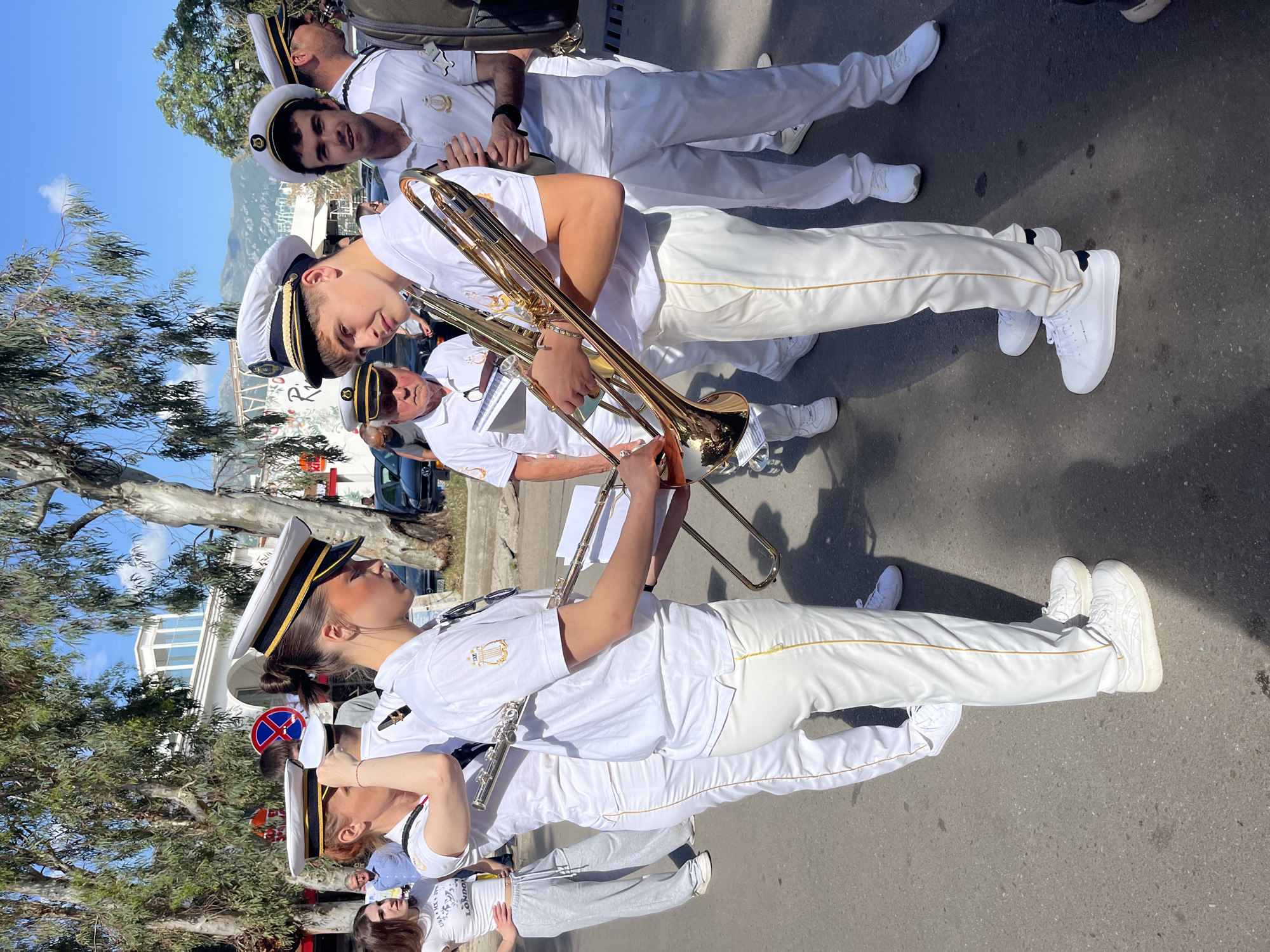 A street-level, eye-level medium shot captures a group of people wearing what appear to be navy uniforms, possibly a marching band, on a sunny day.
The individuals are wearing white uniforms with caps, suggesting a formal event. One person is playing a brass trombone, while another holds a flute. The uniforms include white pants, white shirts with emblems, and caps with dark trim. The people appear to be either standing or walking, likely as part of a parade or performance.
The backdrop shows buildings, trees, and a blue sky, typical of an urban environment. A street sign is visible in the background. The location can be identified as Budva, Montenegro, given the typical vegetation, building style, and overall atmospheric conditions.