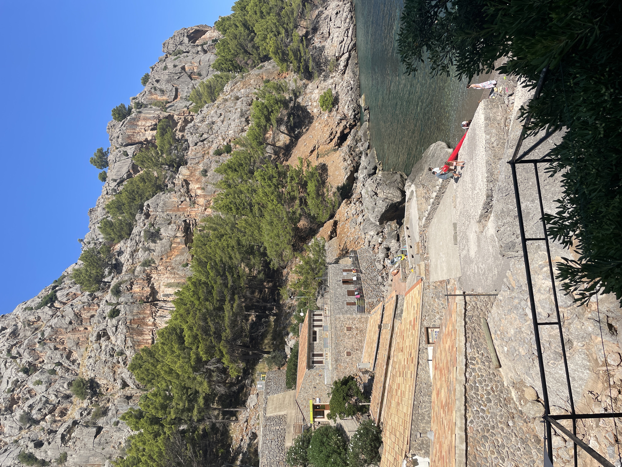 The image depicts a scenic view of Port de Sa Calobra, Spain, on a clear, sunny day. A rugged, tree-lined cliff face dominates the background, sloping down towards a calm, turquoise body of water. Several traditional stone buildings with terracotta tiled roofs are nestled into the hillside, overlooking a small, pebbly beach area.
In the foreground, a group of people are gathered near the water. Some are standing, while others appear to be interacting with a red inflatable object, possibly a raft or kayak, on the shore. A woman in a patterned dress stands apart, looking towards the water. Lush green foliage frames the bottom of the image. The architecture suggests a coastal village setting, with the natural landscape playing a significant role in the overall ambiance.
