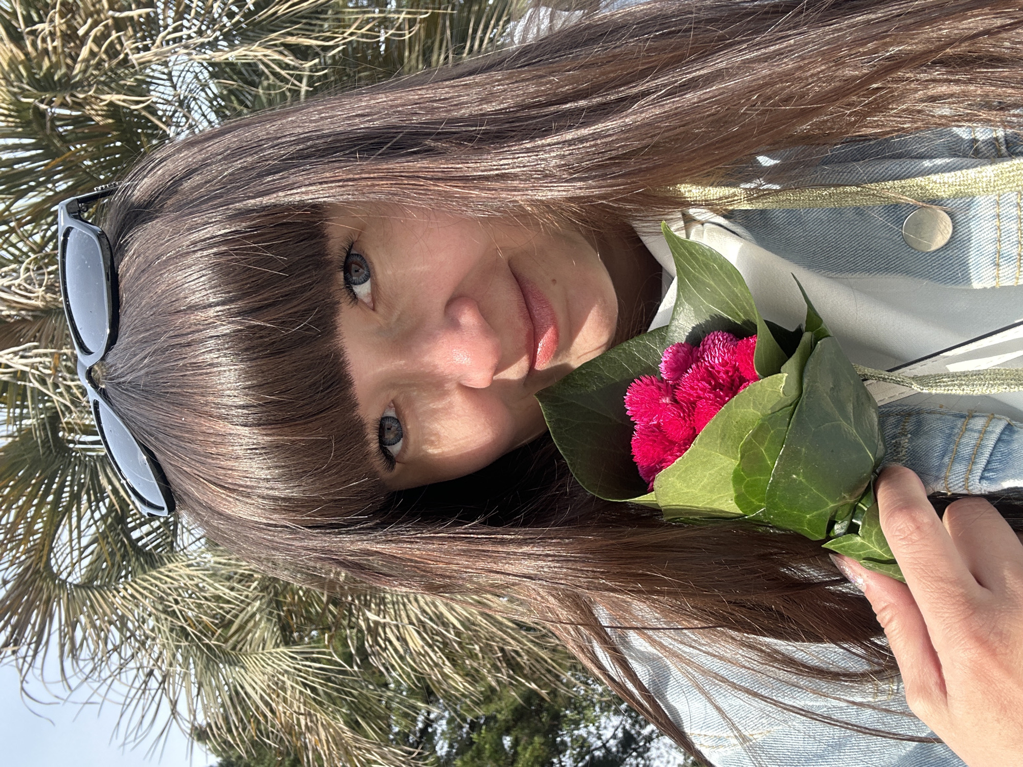 An outdoor portrait captures a young woman from a slightly tilted, close-up perspective, looking towards the viewer with a soft, pleasant expression. She has long, dark brown hair with bangs that frame her blue eyes. Black sunglasses are perched on top of her head.
The woman is wearing a light-colored denim jacket, detailed with a light green trim and a silver-toned button, over a white shirt. She holds a small, vibrant bouquet of bright pink, fluffy flowers, likely Celosia, which are wrapped in large green leaves, in her right hand. Her manicured nails are light and possibly iridescent.
The background features the distinct fronds of palm trees under a bright sky, suggesting a sunny day in a warm climate. This aligns with the provided context of Sochi, Russia, a city known for its subtropical flora. The lighting indicates it is daytime, with clear weather. There is no visible text in the image. The overall mood is casual and serene.