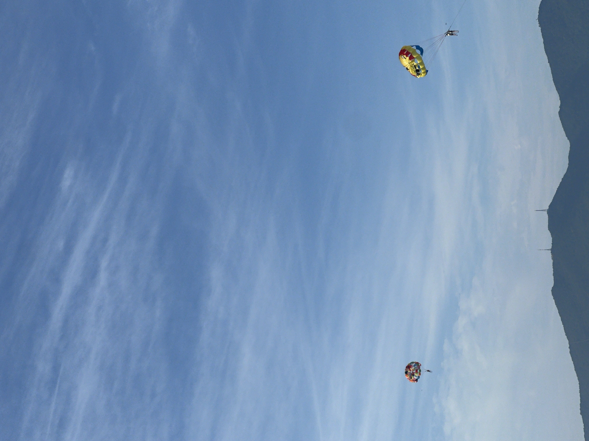 Two parasailing boats are visible in the sky above the coast of Da Nang, Vietnam. The parasail on the left is multicolored with red, orange, yellow, and blue sections. The parasail on the right is yellow with red and blue accents and appears to be shaped like a smiling face. Both parasails have people attached below them, being towed by unseen boats. In the background, a dark mountain range with a few scattered structures is visible. The sky is a clear blue with thin, wispy clouds.