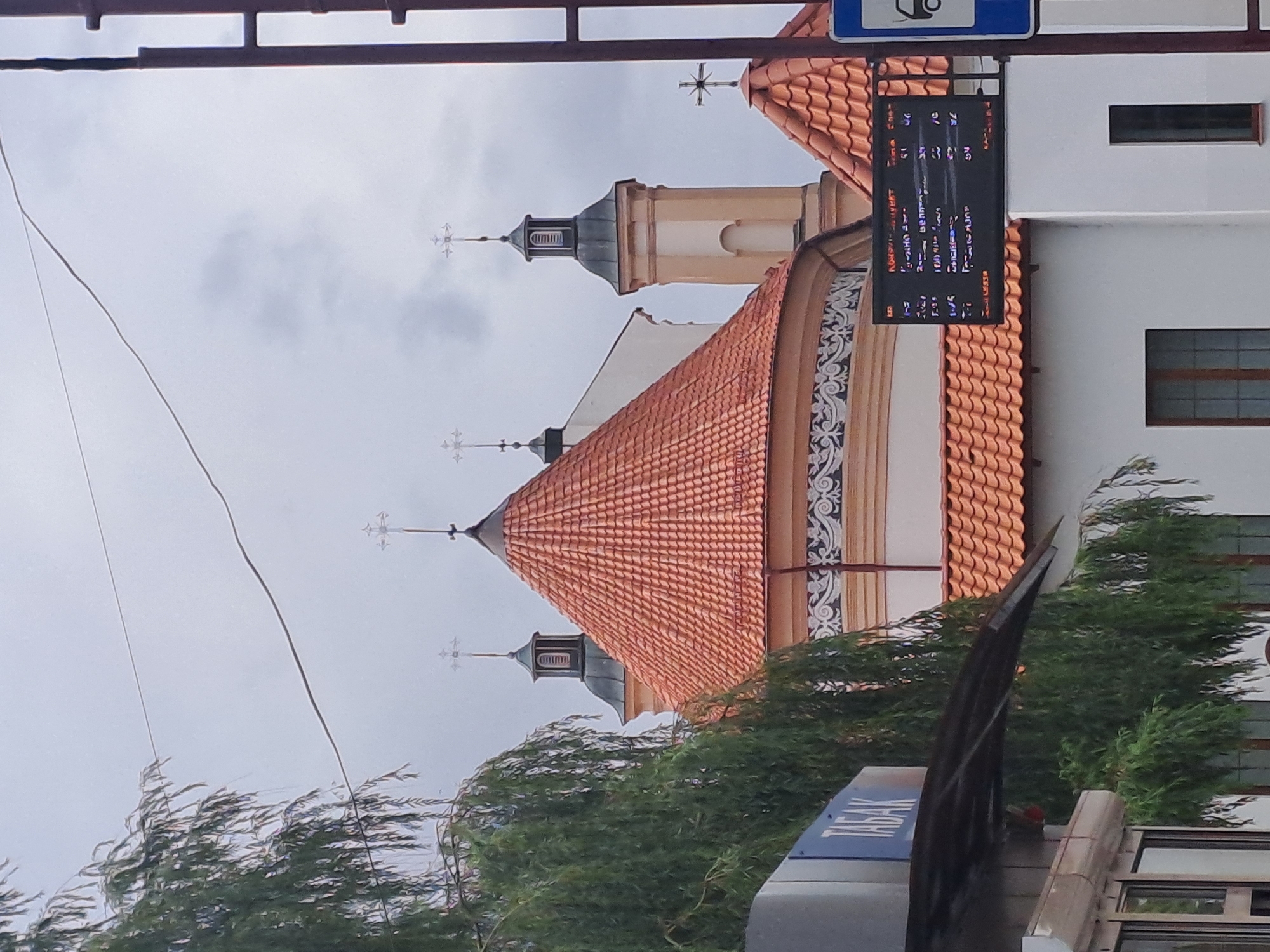 The image shows a view of the Church of Saints Boris and Gleb, also known as Kolozha Church, in Hrodna, Belarus. The church has red tiled roofs and several spires topped with crosses. A portion of a weeping willow tree is visible on the left side of the frame, obscuring some of the church's facade. In the foreground, there is a blue sign with the word "ТАБАК" (tobacco) written on it, suggesting the presence of a tobacco shop. Below the sign is a dark awning. To the right of the church, a digital display board shows what appears to be a schedule or timetable with Cyrillic text and numbers. The sky is overcast, indicating a cloudy day.