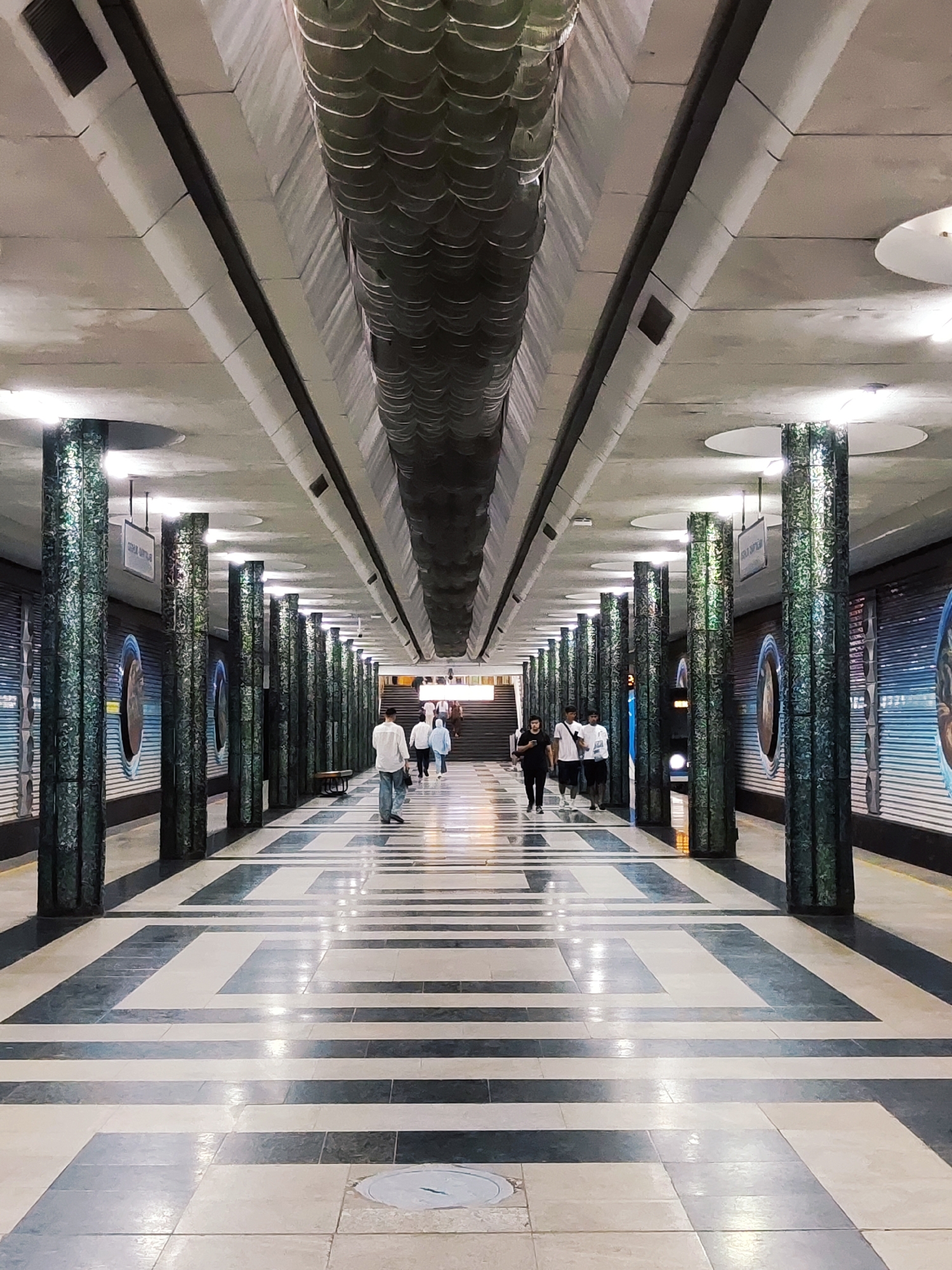 The image shows an interior view of the Tashkent Metro, specifically the Kosmonavtlar metro station in Tashkent, Uzbekistan. The station features a long, wide passageway with a patterned floor made of light and dark grey tiles arranged in geometric shapes. The ceiling is a textured concrete structure with long, curved elements running down the center and embedded lighting.
The passageway is lined on both sides with tall, cylindrical columns that are dark green and textured, appearing to be made of mosaic tiles. Each column is illuminated by spotlights from above. On the left side, behind the columns, are decorative round murals or circular displays with blue and white imagery. On the right side, the wall is also lined with similar circular elements.
Several people are walking through the passageway, appearing to be commuters. On the left side of the walkway, closer to the viewer, a man in a white shirt and blue jeans is walking away from the camera. Further down the passageway, a group of people are walking towards the camera, including individuals in white and light blue clothing. On the right side, closer to the camera, a group of three young men are walking away from the camera, two in white shirts and one in a black t-shirt. In the distance, stairs can be seen leading upwards. The overall impression is a clean, well-lit, and architecturally distinctive metro station.