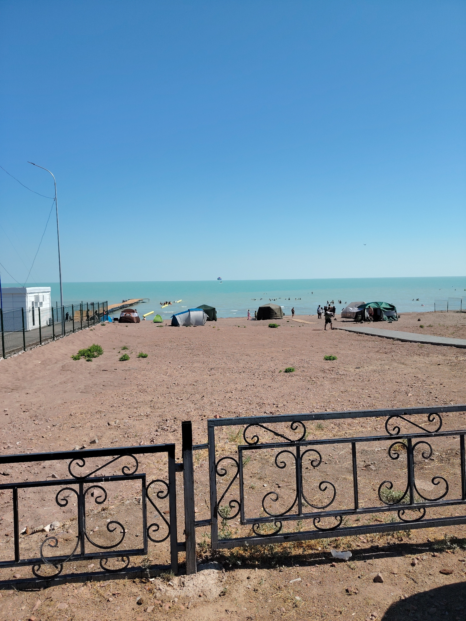 The image depicts a sunny day at a beach in Toranghilik, Kazakhstan. The foreground features a black wrought-iron fence with decorative swirls. Beyond the fence, a sandy beach is visible, with several tents set up. A paved pathway leads towards the water. In the distance, the turquoise water of the sea is calm, with a pier extending into it. Several people are seen swimming and relaxing in the water. Further out, a boat with people on it and a parasailer are visible. A white building with a fence around it is situated on the left side of the image, and a street lamp stands nearby. The sky is clear blue.