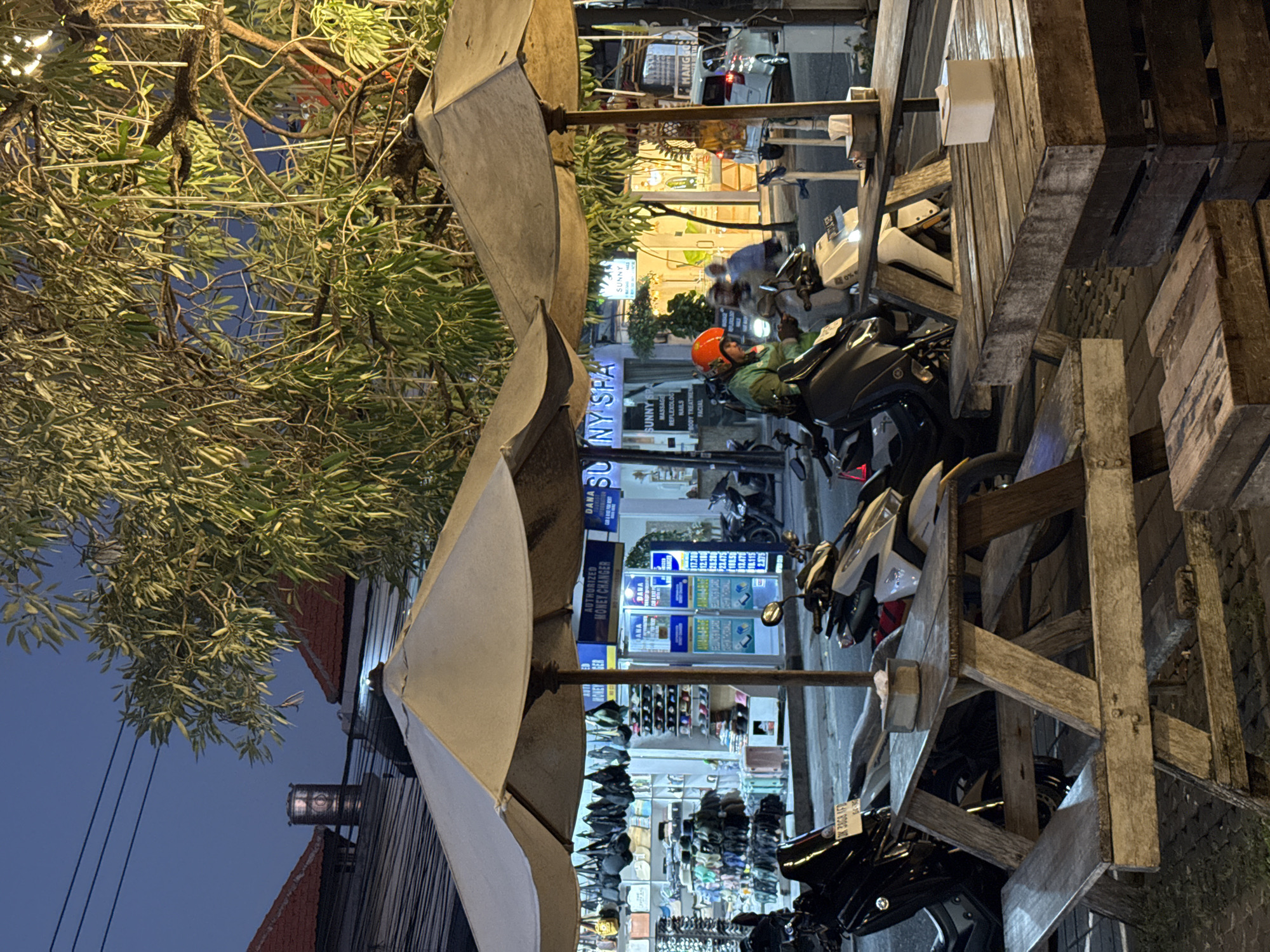 This image depicts a street scene in North Kuta, Indonesia, during the early evening. Two large, light-colored umbrellas provide shade over outdoor seating areas. In the foreground, a weathered wooden picnic table with attached benches is visible, with a small tissue box on its surface. To the right, another wooden table is present, also with a tissue box. Parked motorcycles are in front of the tables.
A person wearing a green shirt and an orange helmet is seated on a motorcycle, looking at a mobile phone. Behind them, other motorcycles and a white car are visible. The background features storefronts, including one labeled "SUNNY SPA" and another appearing to be a money changer with various currency exchange rates displayed. Bags and merchandise are displayed in the shop windows. The street is paved with cobblestones, and overhead power lines are present. Lush green trees with hanging lights contribute to the ambiance. The sky is a deep twilight blue.