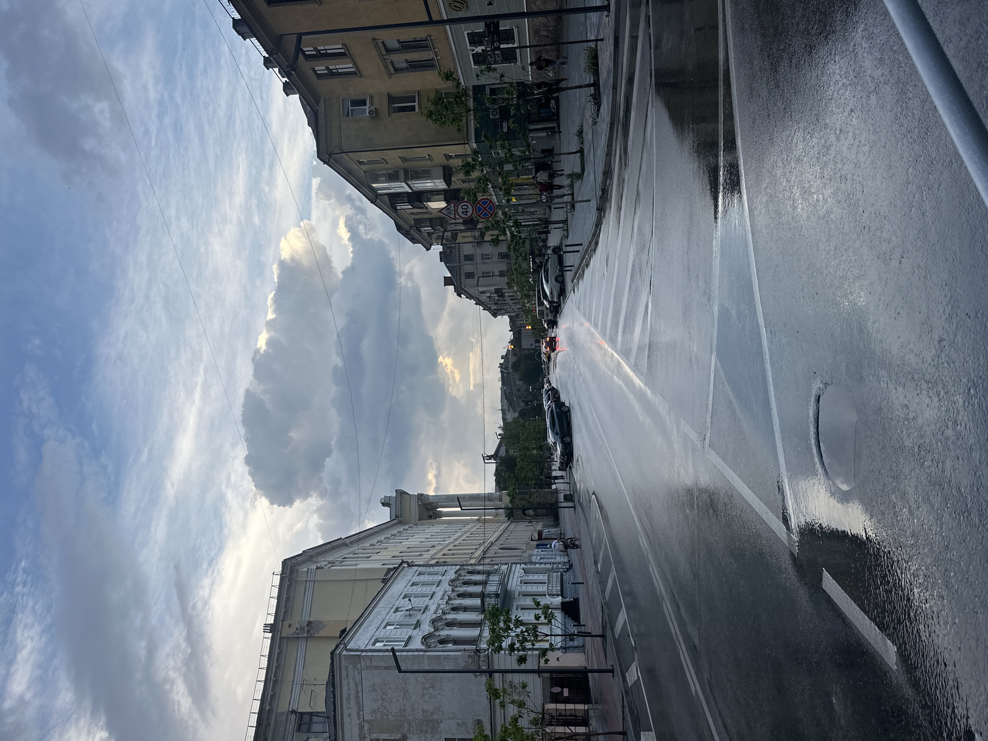 The image depicts a street scene in Ternopil, Ukraine, under a cloudy sky, possibly after a rainfall, as the road surface appears wet and reflective. The street is lined with multi-story buildings, characterized by their architectural styles, suggesting an urban setting. On the left side of the street, a building with classical architectural elements, including columns and ornate window frames, is visible. On the right side, buildings with more contemporary facades, some featuring balconies and air conditioning units, are present. Several cars are parked along the street and some are in motion. A few pedestrians are walking on the sidewalk, and a person is seen riding a bicycle. Traffic signs, including a speed limit sign of 40, are visible. The overall impression is of a somewhat overcast day in a European city, with signs of recent precipitation.