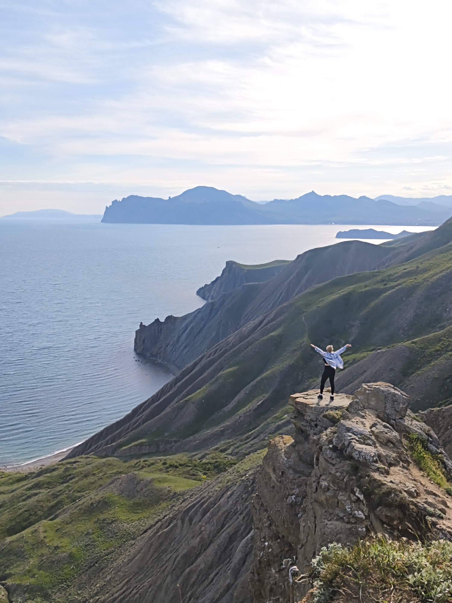 A high-angle, long shot captures a woman standing atop a rocky peak, arms outstretched, with an expansive view of the landscape. The woman is wearing dark leggings, a top, sneakers, and a light-colored jacket. The landscape consists of a coastline with calm ocean waters, rugged mountains, and steep cliffs. The sky is light blue with scattered clouds. The foreground and middle ground showcase the rocky terrain of the mountainside with sparse green vegetation. The horizon is marked by distant landforms. The scene is located in Орджоникидзевский поселковый совет, Ukraine.