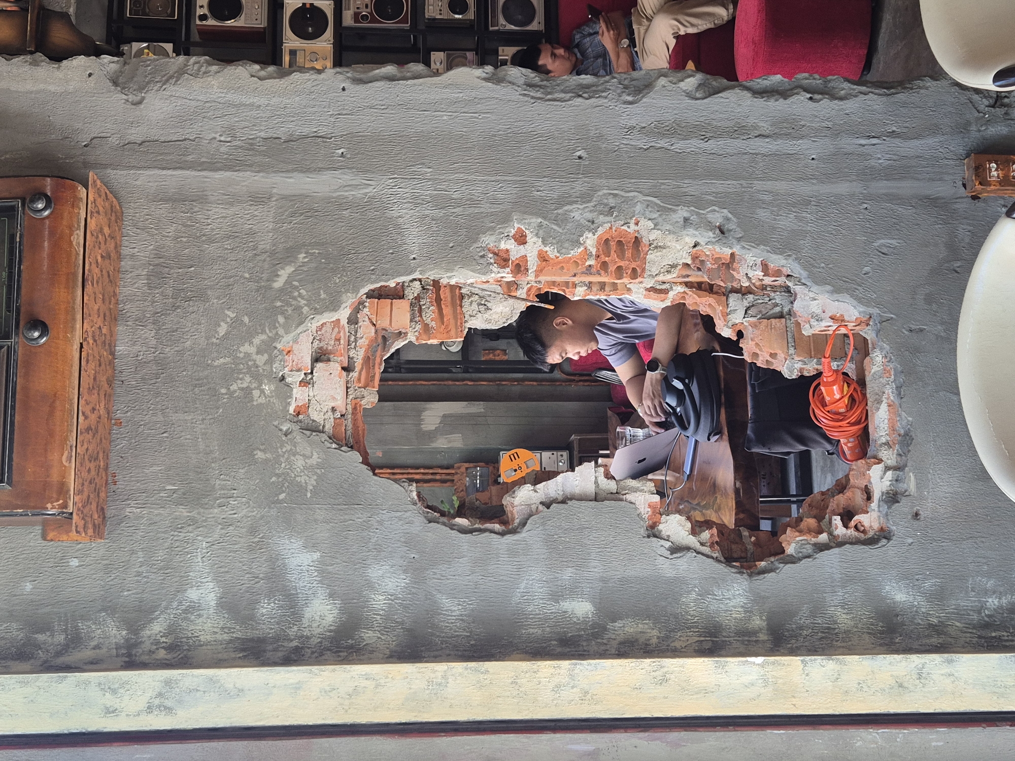 This image depicts a cafe in Da Nang, Vietnam, characterized by its raw, industrial aesthetic. A prominent feature is a large, irregular hole in a concrete wall, revealing exposed red bricks and internal plumbing. Through this opening, two men are visible. In the foreground, a man sits at a wooden table, looking at a laptop, with a glass of water and a black bag nearby. His left hand is on the keyboard, and his right is near a black steering wheel-like object, possibly for a VR headset. To the right, another man sits on a red sofa, looking at his phone. The background displays shelves filled with vintage-looking radios and speakers, adding to the cafe's retro vibe. The lighting suggests daytime.