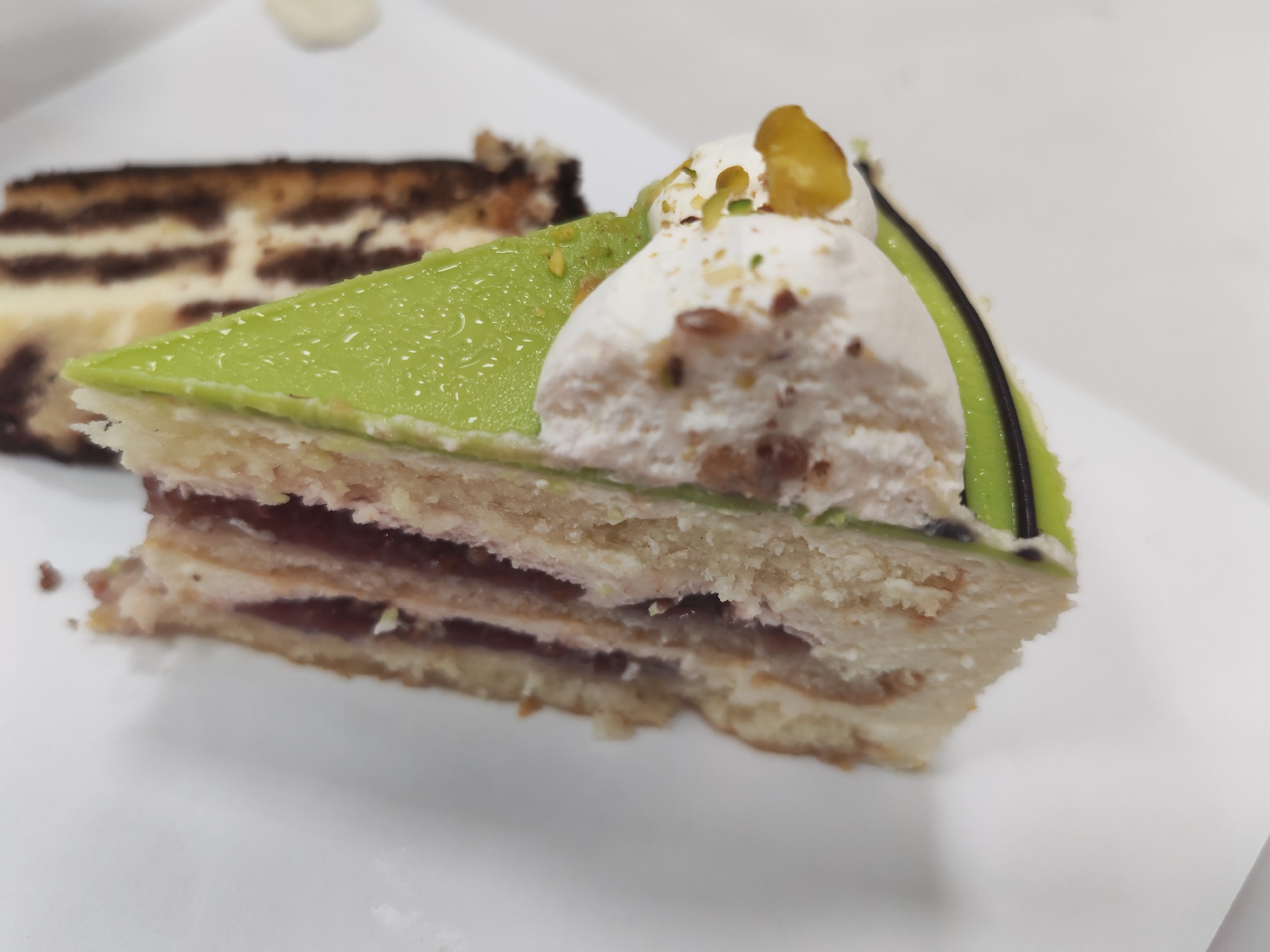 A close-up shot shows two slices of cake on a white surface. The slice in the foreground is a triangular piece with layers of white cake, a red filling, and a bright green frosting on top. It is adorned with white cream, chopped nuts, and a yellow garnish. A dark green line runs along the edge of the green frosting. The second slice of cake, slightly out of focus in the background, appears to be a chocolate and vanilla marbled cake with cream filling. The overall scene suggests a dessert display or a cafe setting. The image's location in Izhevsk, Russia, is not discernible from the visual elements.