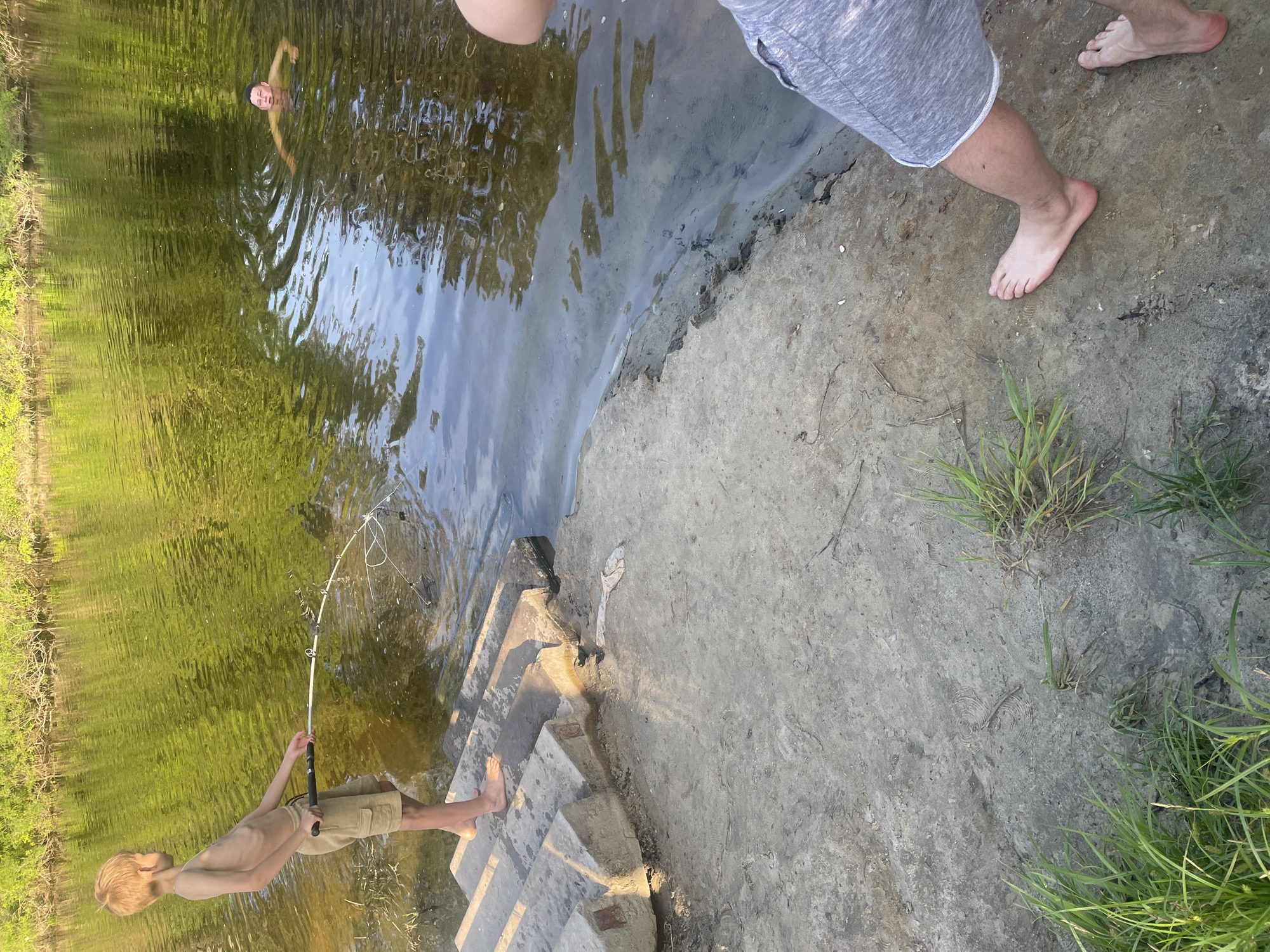 This image captures an outdoor scene at a river or large stream in Petrivka, Ukraine, under bright daylight conditions.
Three individuals are present. On the left, a fair-skinned boy, approximately 10-14 years old, with short blonde hair, stands shirtless and barefoot on the lowest of four concrete steps that descend into the water. He wears light brown cargo shorts. The boy is holding a white fishing rod, the tip of which is bent downward, indicating tension. A tangled, white, net-like or debris object is visible at the end of the fishing line, partially submerged in the shallow water directly in front of him.
Further out in the greenish-brown water, an adult or older teenager with dark hair is partially submerged, with only their head and shoulders visible above the water, appearing to be floating or treading water on their back. Their face shows a discernible expression.
In the lower right foreground, the lower torso and legs of a third individual, an adult, are visible. This person is standing barefoot on the bare, light grey-brown earthen bank, wearing grey shorts.
The body of water displays a greenish-brown coloration with visible surface ripples and reflections of the sky and foliage. The far bank is densely covered with lush green trees and bushes. The immediate foreground consists of compacted earth with scattered patches of green grass and small debris. The concrete steps are worn and partially submerged where they meet the water. Shadows are cast by the steps and individuals, suggesting the sun is at a moderate angle, likely in the late afternoon.