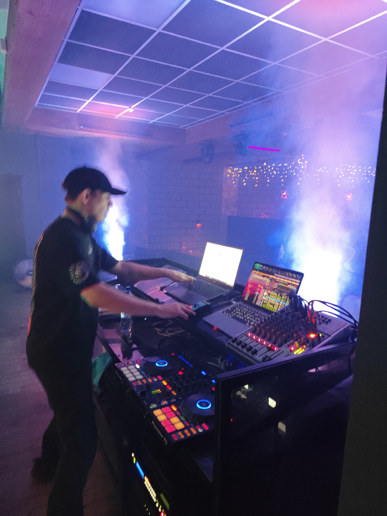 The image depicts a DJ in a nightclub setting, presumably in Chaikovsky, Russia, although this is not definitively confirmed. The DJ, wearing a black t-shirt and baseball cap, is operating electronic equipment, including a Pioneer DJ controller, a mixer, and two laptops. One laptop screen displays audio waveforms, indicating music production or mixing software. The other is blank. The equipment is set up on a stand. The environment is dimly lit with blue and purple stage lighting and artificial smoke. String lights are visible in the background. The ceiling features a grid-like design.