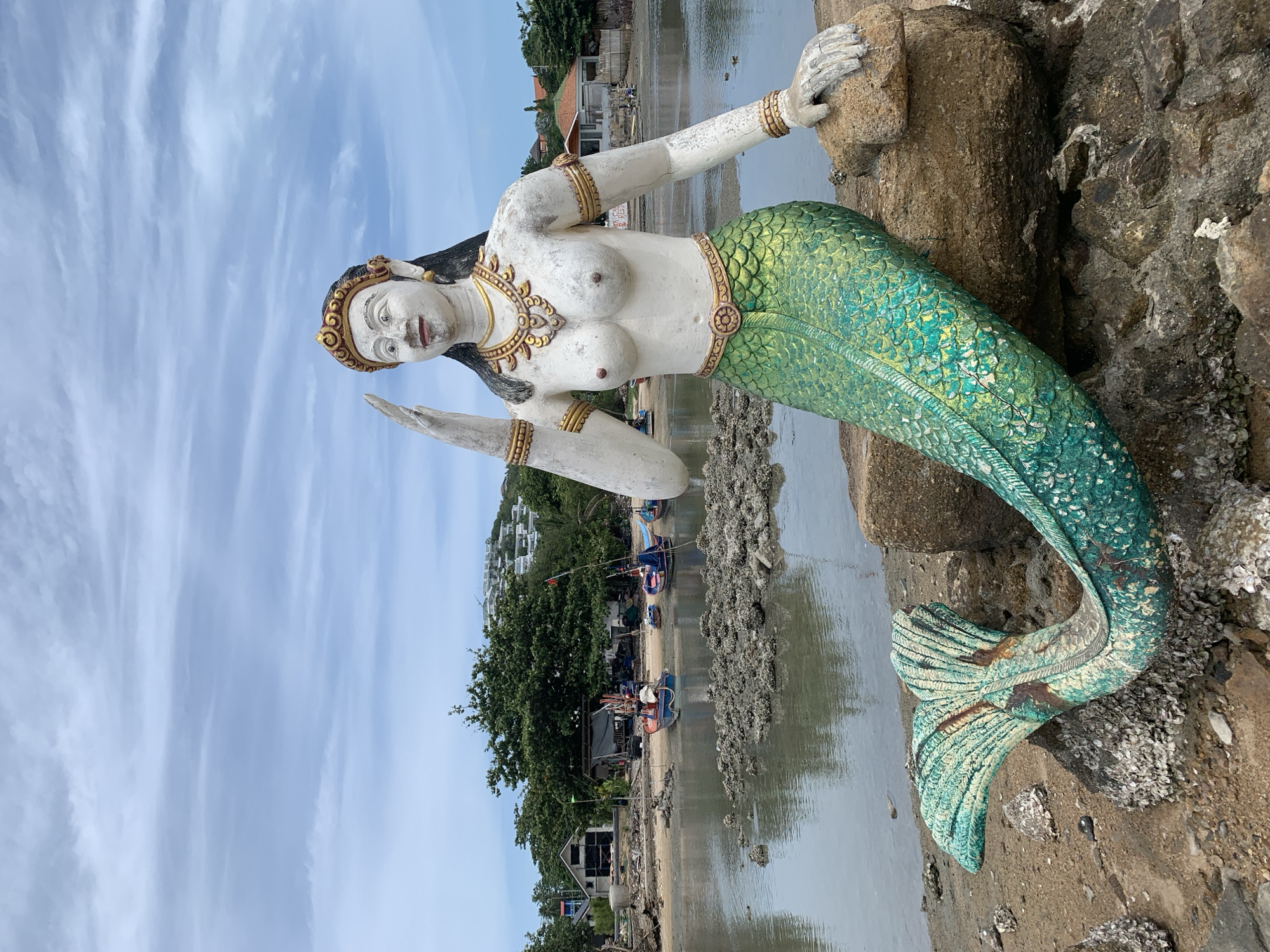 A full-body, weathered statue of a mermaid sits on rocks at the edge of the water in Ko Samui District, Thailand. The statue, painted white with gold accents and a green, scaly tail, has its right hand raised in a wave. Behind the statue, a shallow, murky body of water with scattered rocks leads to a sandy shore. Several small, colorful fishing boats are pulled up on the shore, adjacent to simple structures and lush green trees. In the distance, the hazy outline of buildings on a hillside is visible under a cloudy sky. The overall scene depicts a coastal area with a touch of local artistry.