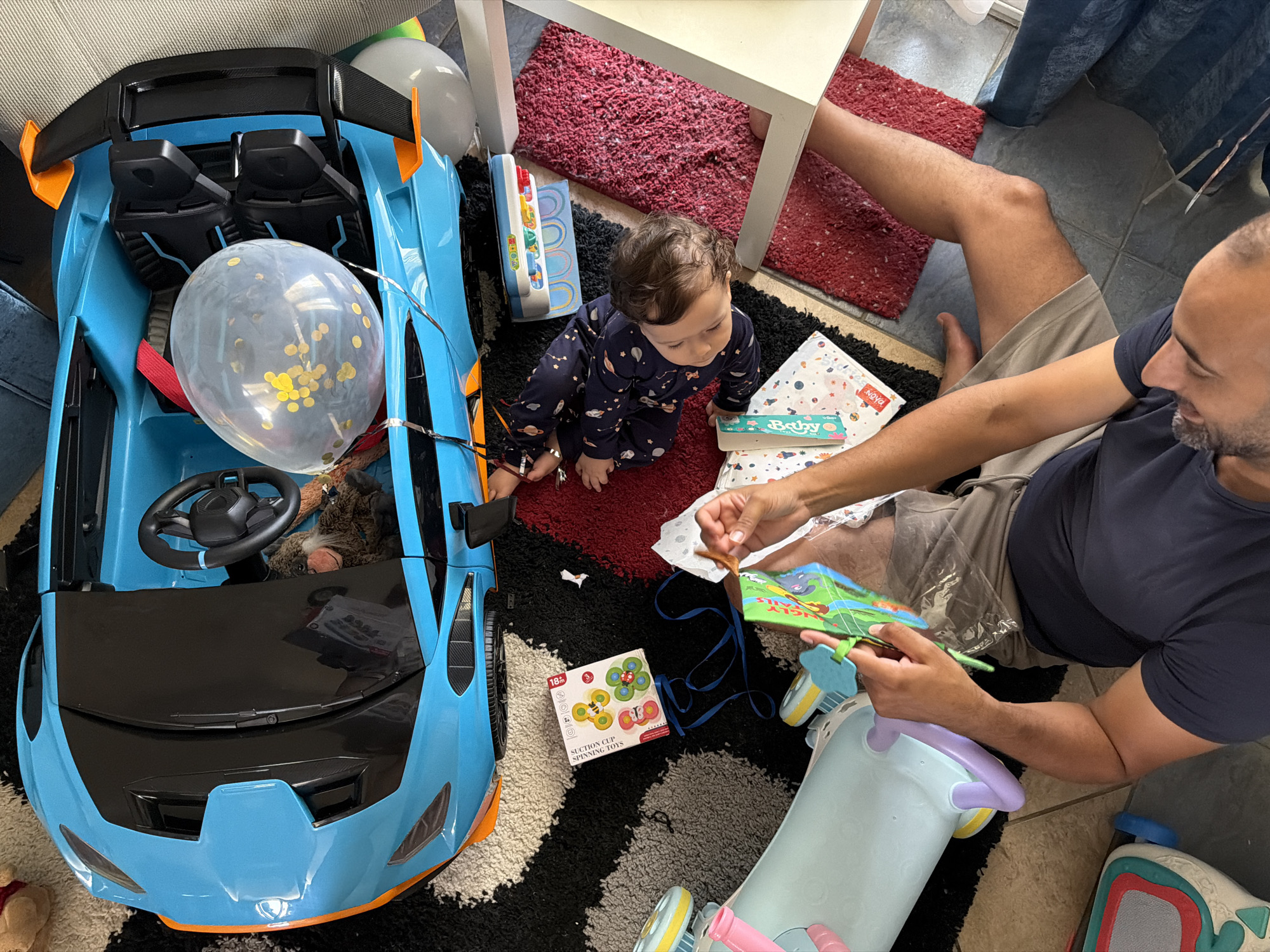 The image depicts a bright, indoor scene, likely a child's playroom or living area. A large, blue toy car with orange accents is the central focus. A baby, dressed in a dark blue outfit with white stars, sits on a red rug next to the car. The baby is looking down at a book in front of them. A man, wearing a dark blue t-shirt and light grey shorts, is also on the floor, holding a green book with colorful illustrations. He appears to be interacting with the book, possibly reading it to the baby. A translucent balloon with gold confetti floats inside the toy car. Scattered around the scene are various other toys, including a pale blue push toy with purple accents, a colorful toy keyboard, and a box labeled "Spinning Toys." The floor is covered with a patterned rug in black, white, and grey, with a separate red rug visible. The overall impression is one of a playful and domestic setting.