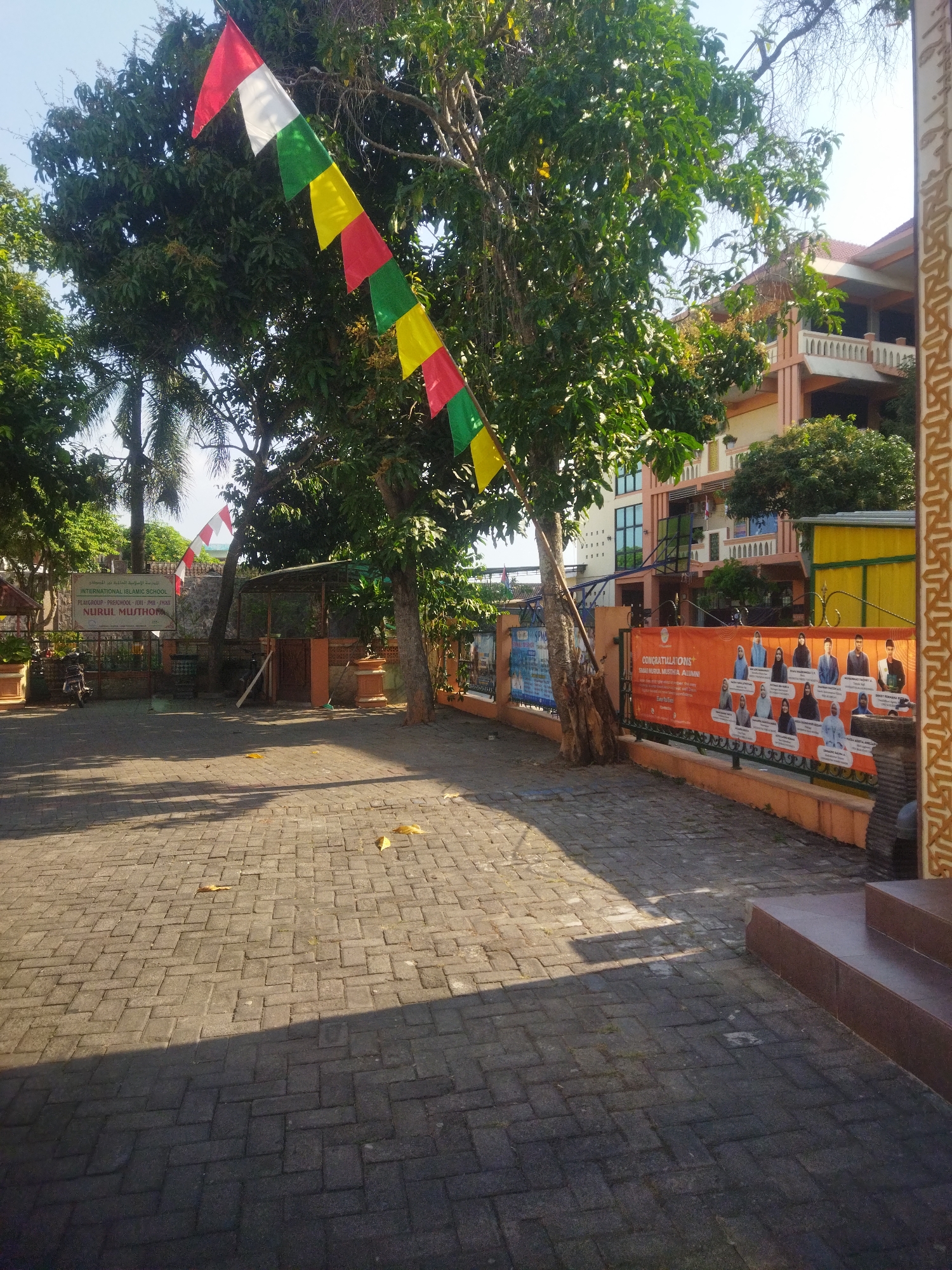 The image shows a paved courtyard with a brick walkway leading to stairs on the right. Several trees with green foliage are present, one of which is decorated with a string of red, white, green, and yellow flags. In the background, a building with multiple stories and balconies is visible, as well as a sign that reads "INTERNATIONAL ISLAMIC SCHOOL NURUL MUTTHOHAROH". Another banner, orange in color, displays portraits of individuals and the text "CONGRATULATIONS SMA NURUL MUTTHOHAROH ALUMNI". The scene appears to be a school campus in Juwiring, Indonesia. The lighting suggests it is daytime, with shadows cast across the walkway.