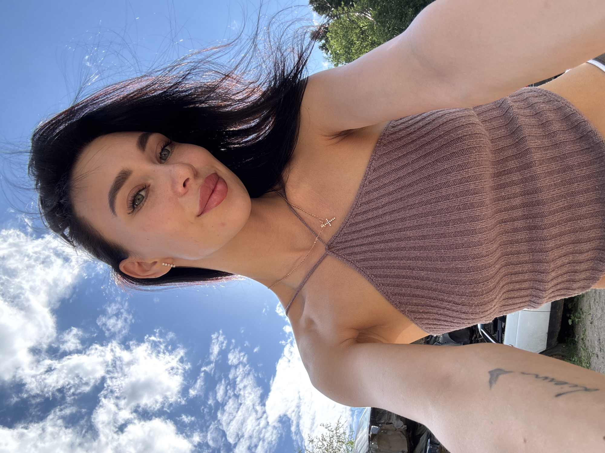 The image is a selfie taken outdoors on a sunny day. The background consists of a bright blue sky with scattered white clouds, green trees on the right, and a pile of debris and a white vehicle on the left. The subject is a young woman with long, dark hair, fair skin, and green eyes. She is wearing a knitted halter top with a ribbed texture. A gold cross necklace is visible around her neck. She has a tattoo on her left forearm. The location is the city of Chaikovsky, Russia.