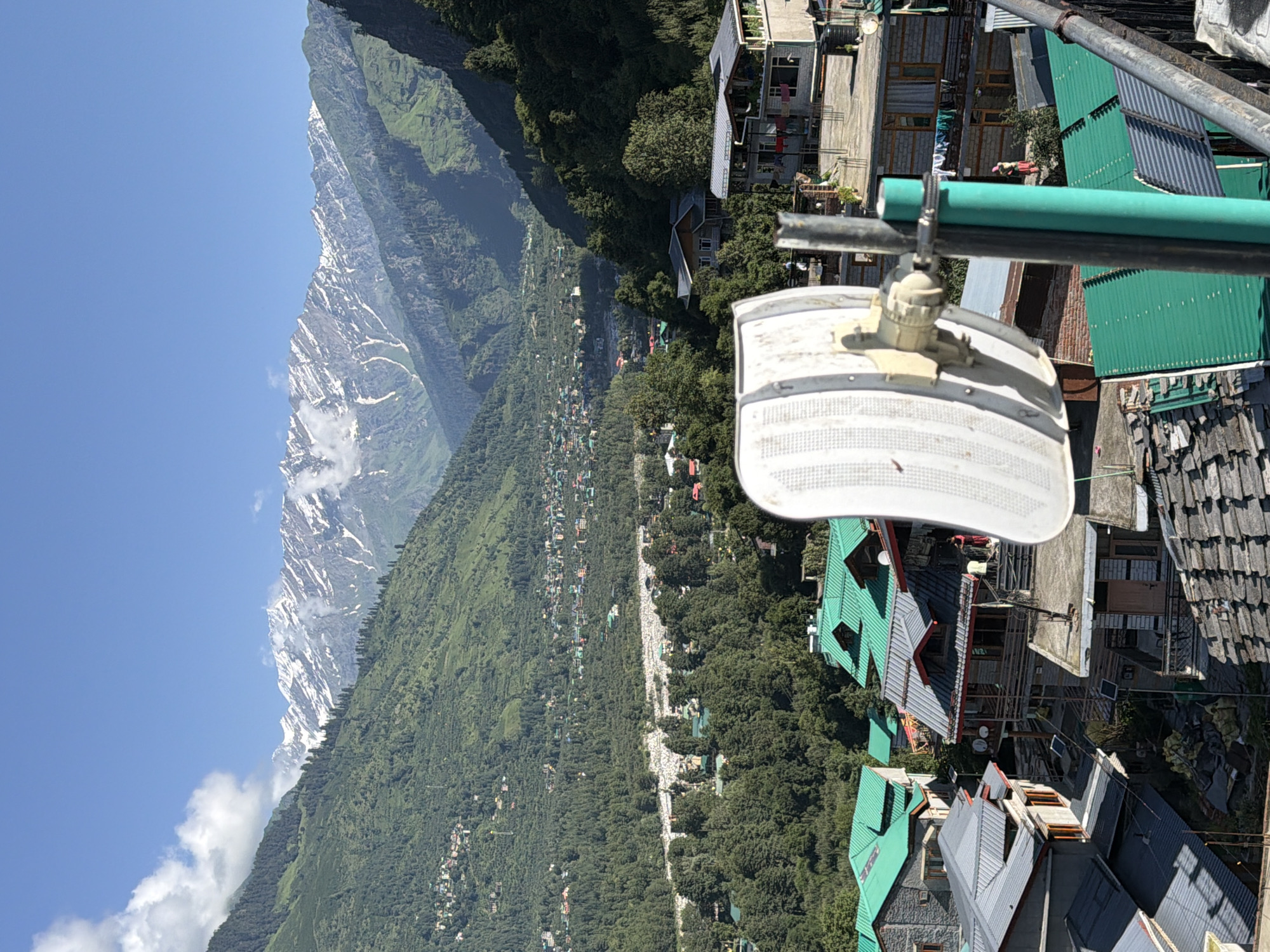 A high-angle outdoor shot captures a scenic mountainous landscape in Vashisht, India. In the foreground, a white parabolic antenna is mounted on a pole, partially obscuring a cluster of buildings with green metal roofs. The buildings appear to be multi-story structures with traditional architecture. In the middle ground, a valley unfolds, dotted with more buildings and dense green foliage. The background is dominated by a majestic mountain range, with snow-capped peaks piercing a clear blue sky. The overall scene conveys a sense of natural beauty and rural life.