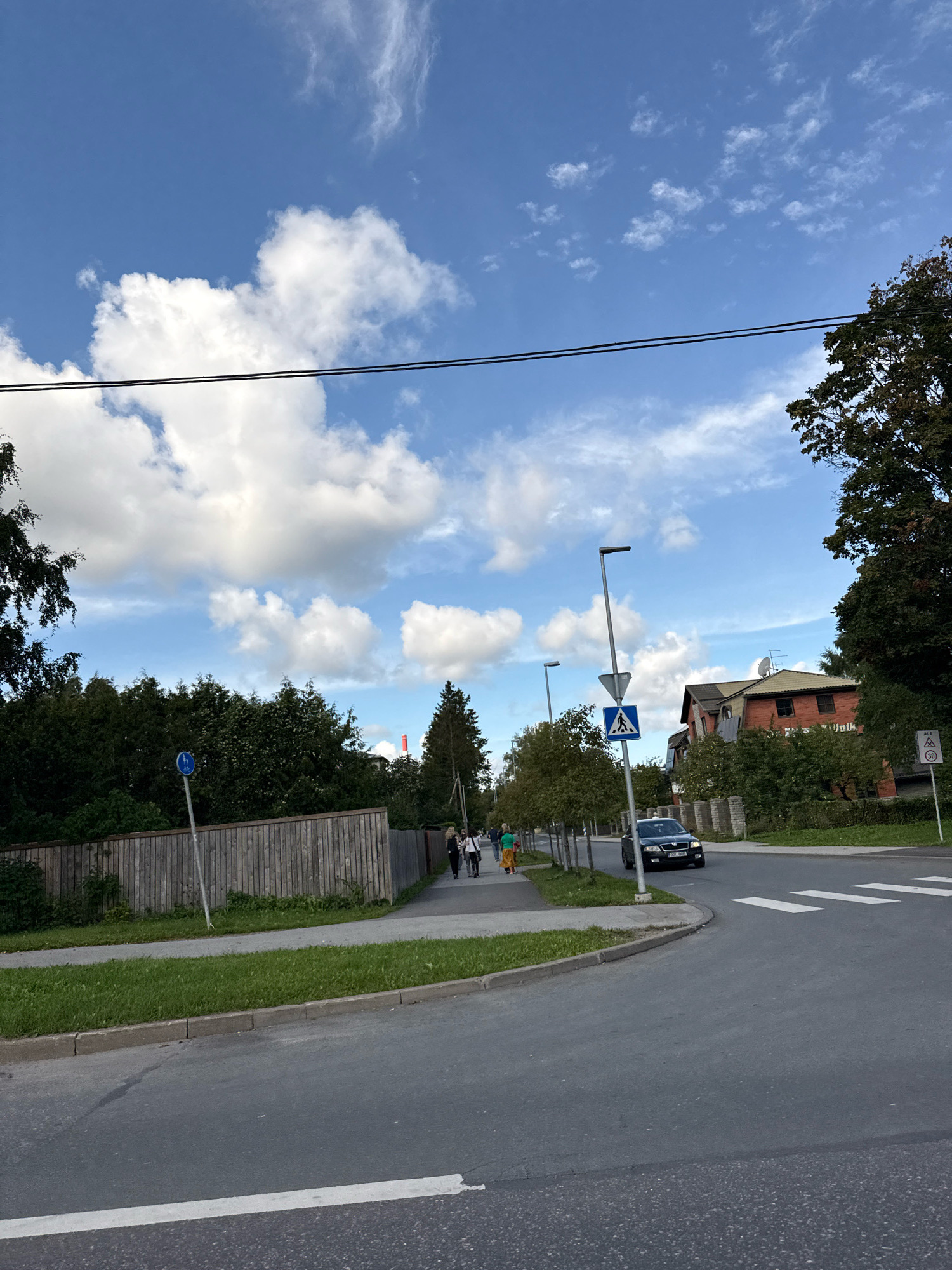 The image captures a street scene in Tallinn, Estonia, under a bright blue sky with scattered white clouds. A paved road curves to the right, with a white pedestrian crossing marked on it. To the left of the road, a sidewalk is bordered by a grassy verge and a wooden fence. A tall street lamp stands on the sidewalk, with a pedestrian crossing sign below it. Further down the sidewalk, a group of people are walking, and a dark-colored car is driving on the road. Behind the sidewalk and fence, lush green trees and vegetation are visible, with residential buildings peeking through the foliage in the distance. The overall impression is a tranquil, everyday street scene in a residential area.
