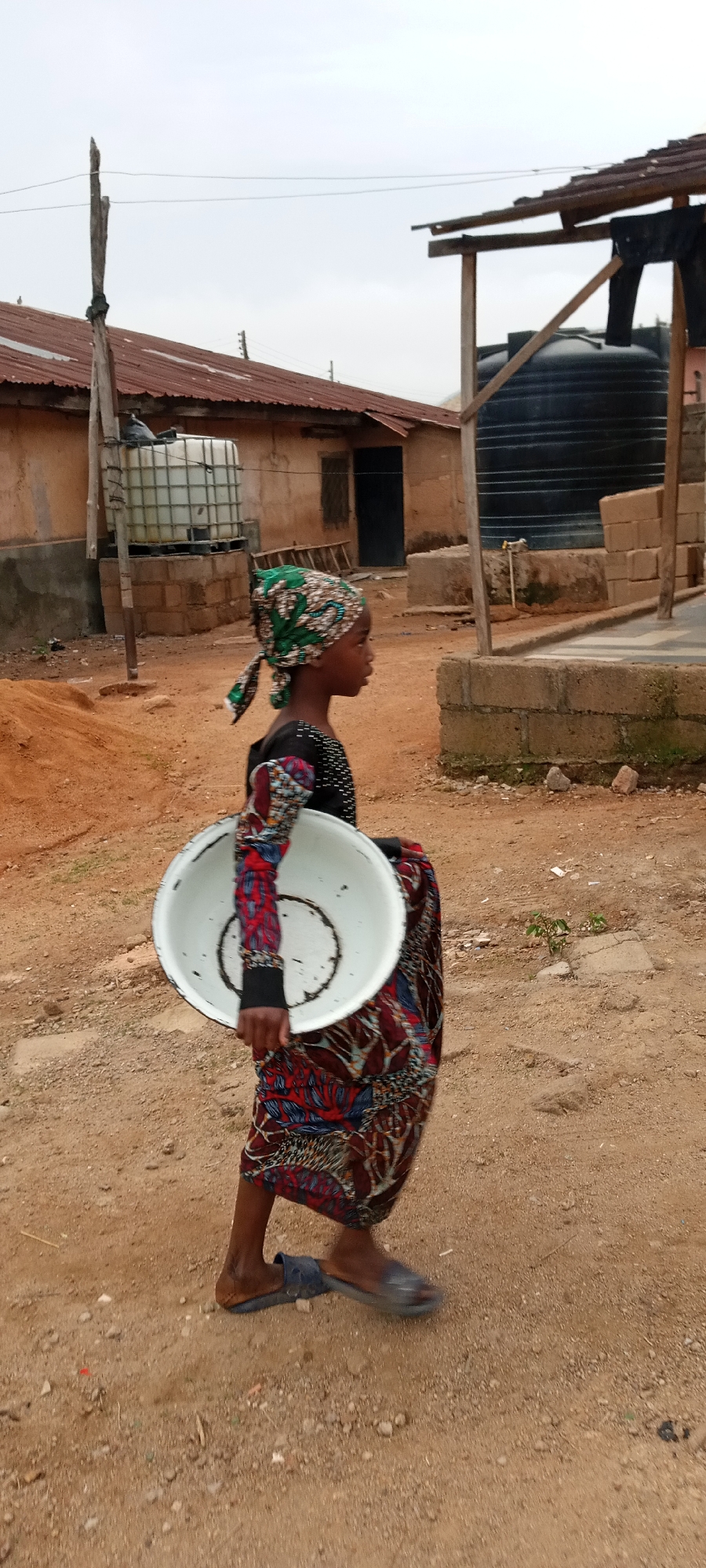A young girl with a green patterned headscarf walks on a reddish-brown dirt road in Jos, Nigeria. She is wearing a long-sleeved black top and a patterned skirt in shades of red, blue, and brown. She carries a large, white, slightly worn bowl in her left hand. The girl is wearing blue flip-flops. In the background are structures typical of a developing area, including buildings with corrugated iron roofs, water tanks (one white and caged, the other black), and simple brick or concrete structures. There is a small pile of red dirt near the building to the left. The sky is overcast.