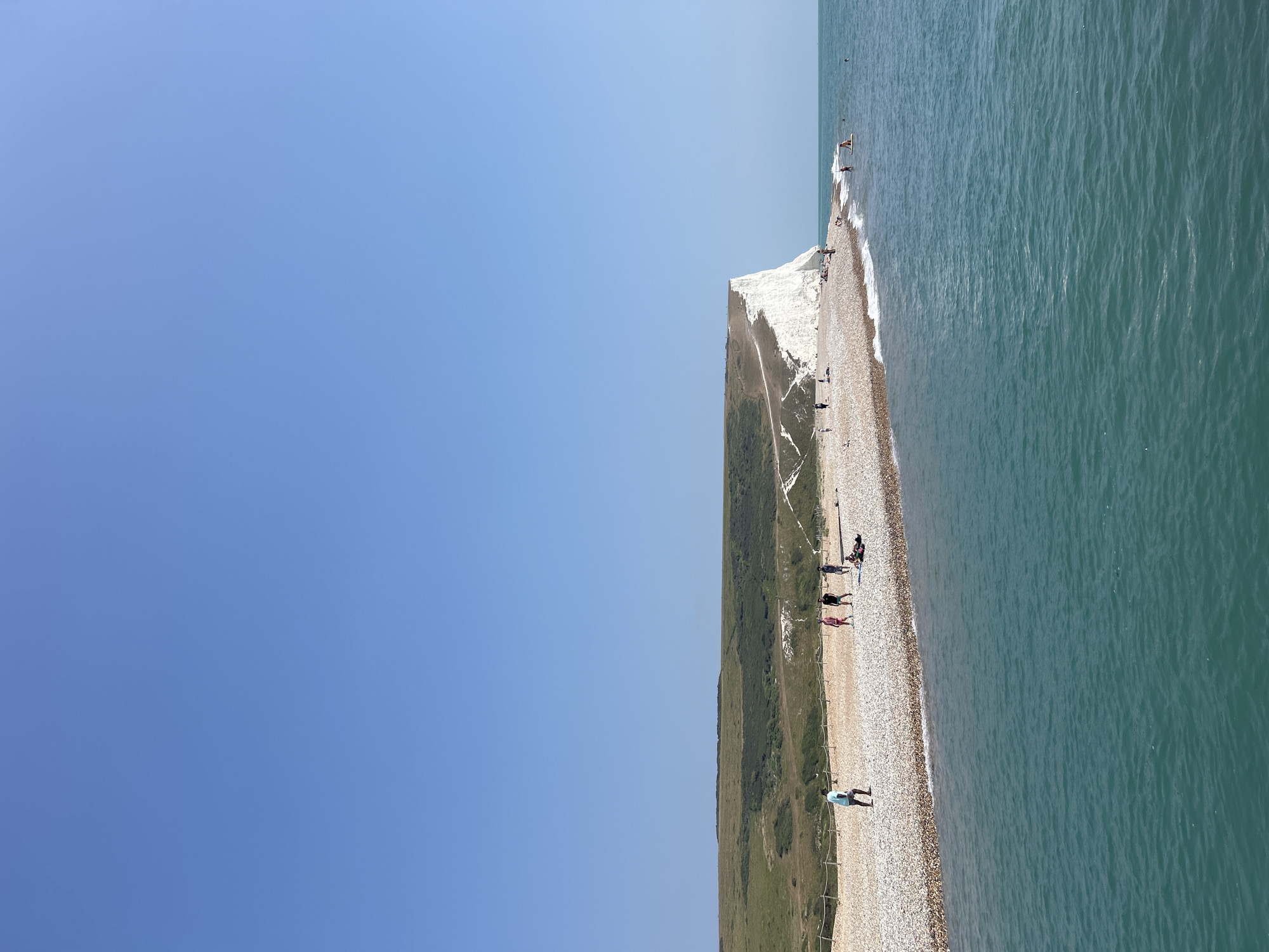 This is a photograph of the Seven Sisters cliffs in Seaford, United Kingdom, on a clear, sunny day. The image is taken from a low angle, looking towards the sea. In the foreground, the turquoise water is calm with small waves lapping at the shore. The beach is made of pebbles, and a few people are visible walking along it or sitting on the shore. Further back, the iconic white chalk cliffs rise dramatically, meeting a green, rolling hillside. The sky above is a vast expanse of clear blue. The scene captures a typical summer day at this popular coastal location.