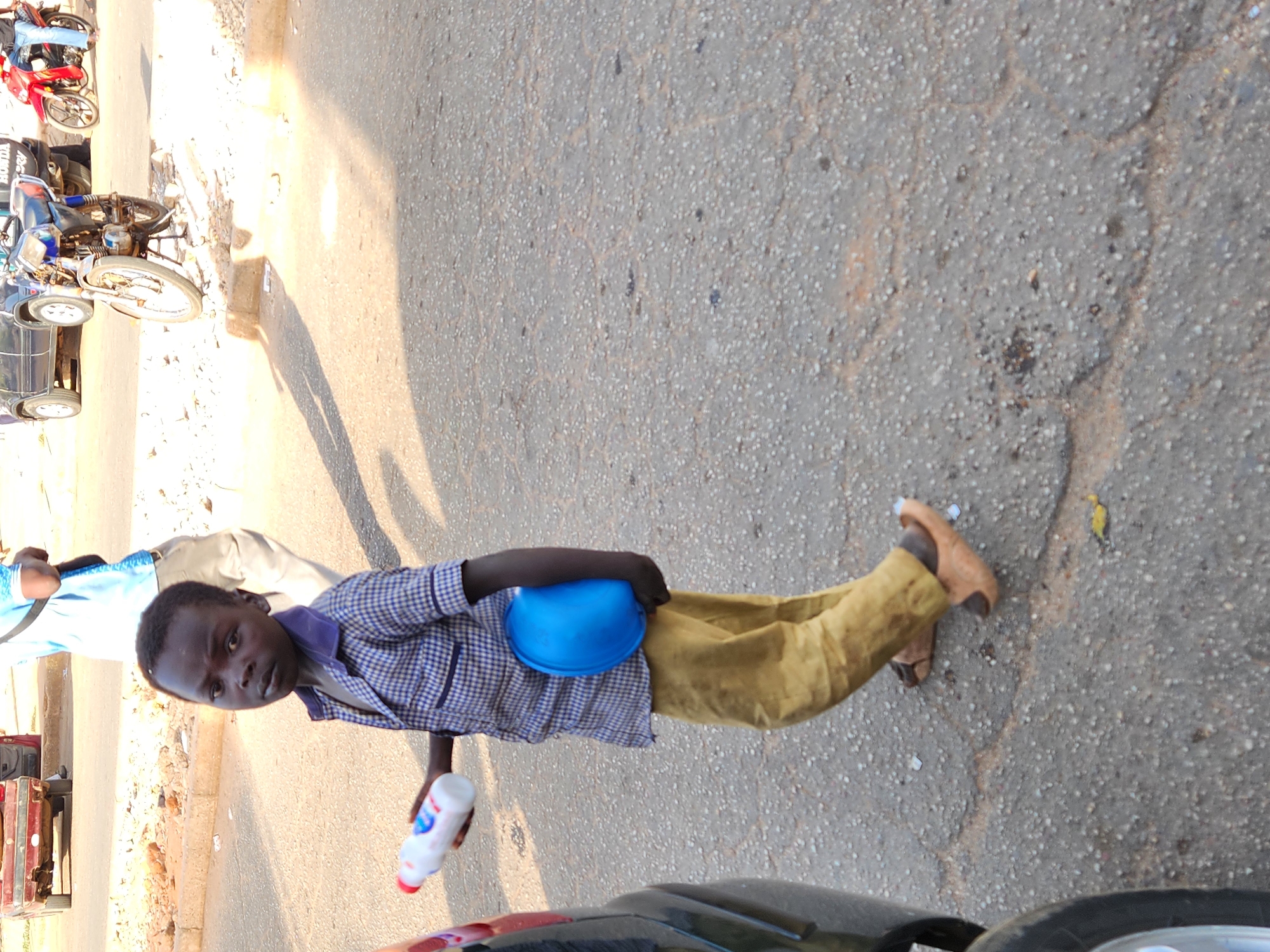 A young boy stands on a paved surface holding a plastic bottle and a blue hard hat. He wears a plaid shirt and tan pants. The location appears to be an urban setting with vehicles and motorcycles visible in the background. The setting is likely Jos, Nigeria.