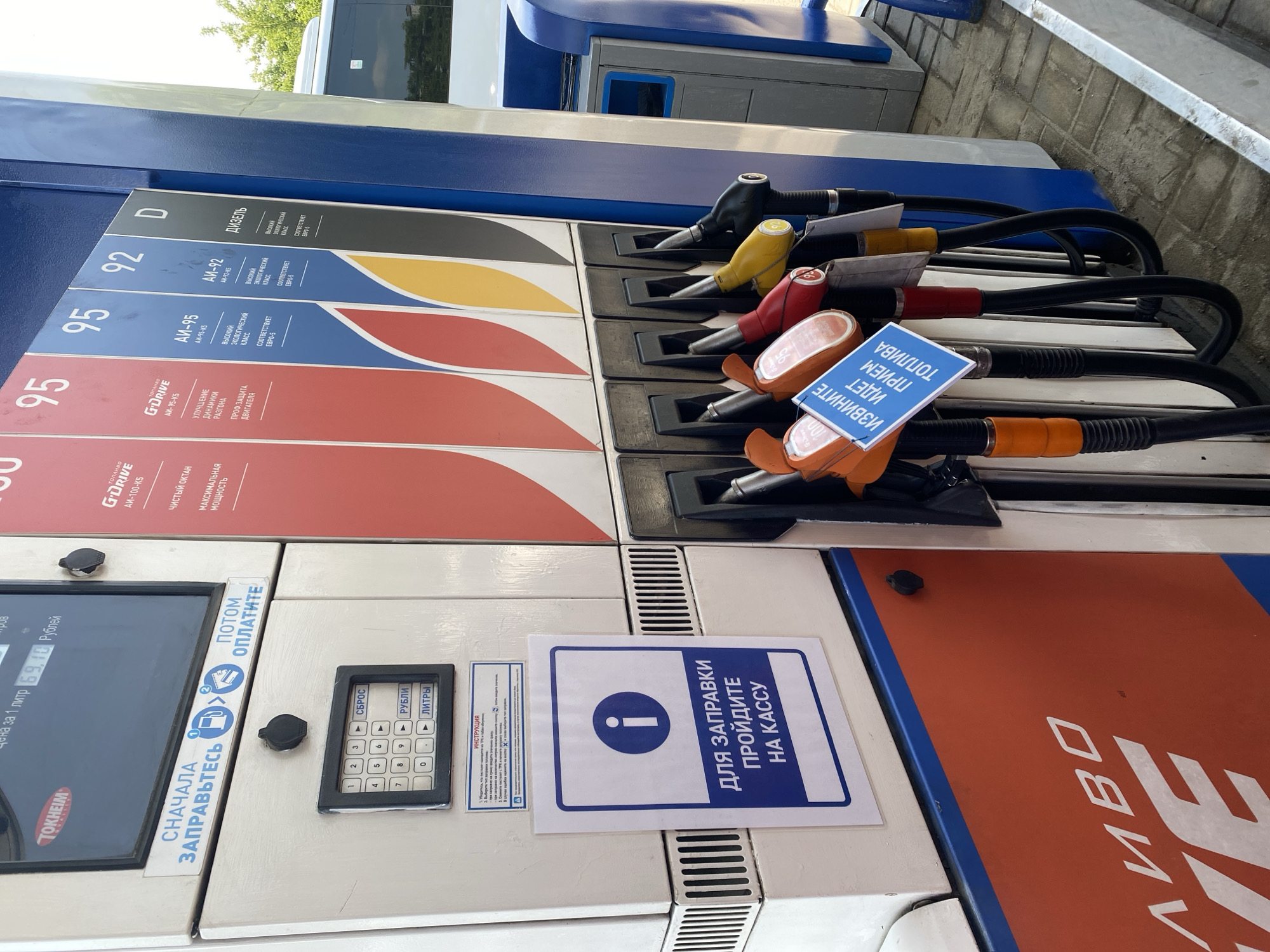 This is a medium-shot, eye-level, outdoor photograph taken at a gas station. The scene focuses on a fuel pump and its dispensers. The pump is white and cream-colored with a black keypad for entering amounts and selecting currency or volume. Above the keypad, a screen displays "цена за 1 литр 69,10 Рублей" (price per liter 69.10 rubles). To the left of the screen, a sign in Russian reads "СНАЧАЛА ЗАПРАВЬТЕСЬ ПОТОМ ОПЛАТИТЕ" (First refuel, then pay) with icons indicating the process. A blue and white sign with an information icon states "ДЛЯ ЗАПРАВКИ ПРОЙДИТЕ НА КАССУ" (For refueling, go to the cashier).
To the right of the pump, a row of fuel dispensers is visible, each with a nozzle and a colored handle. The dispensers are labeled with fuel types: 95, 95, 92, and D (Diesel). Some of the dispensers have small blue and white signs attached, with one clearly visible reading "ИЗВИНИТЕ ИДЕТ ПРИЕМ ТОПЛИВА" (Sorry, fuel delivery is in progress). The background shows a white bus or vehicle and some greenery. The ground is paved with grey concrete. The photograph appears to be taken in Novokuznetsk, Russia, based on the visual cues. No people are visible in the frame, and no actions are actively occurring beyond the static display of the gas station equipment.